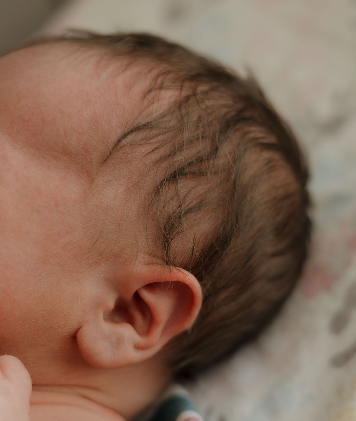 Close-up of a sleeping newborn baby with dark hair and a tiny ear, resting peacefully on a soft blanket.