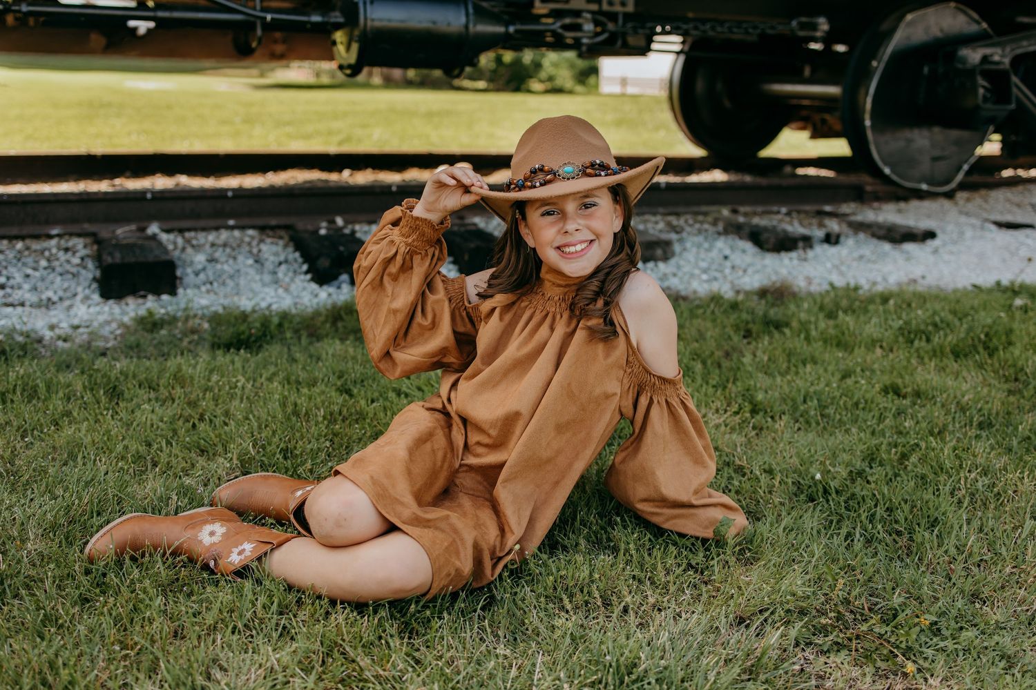 Young girl in brown dress, cowboy hat, and boots sits on grass near railroad tracks, smiling and holding hat.
