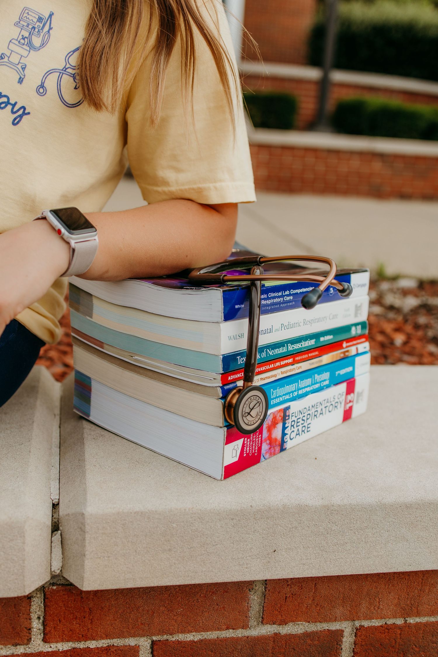 Medical student holding stack of colorful textbooks with stethoscope outdoors, wearing yellow shirt and smartwatch.