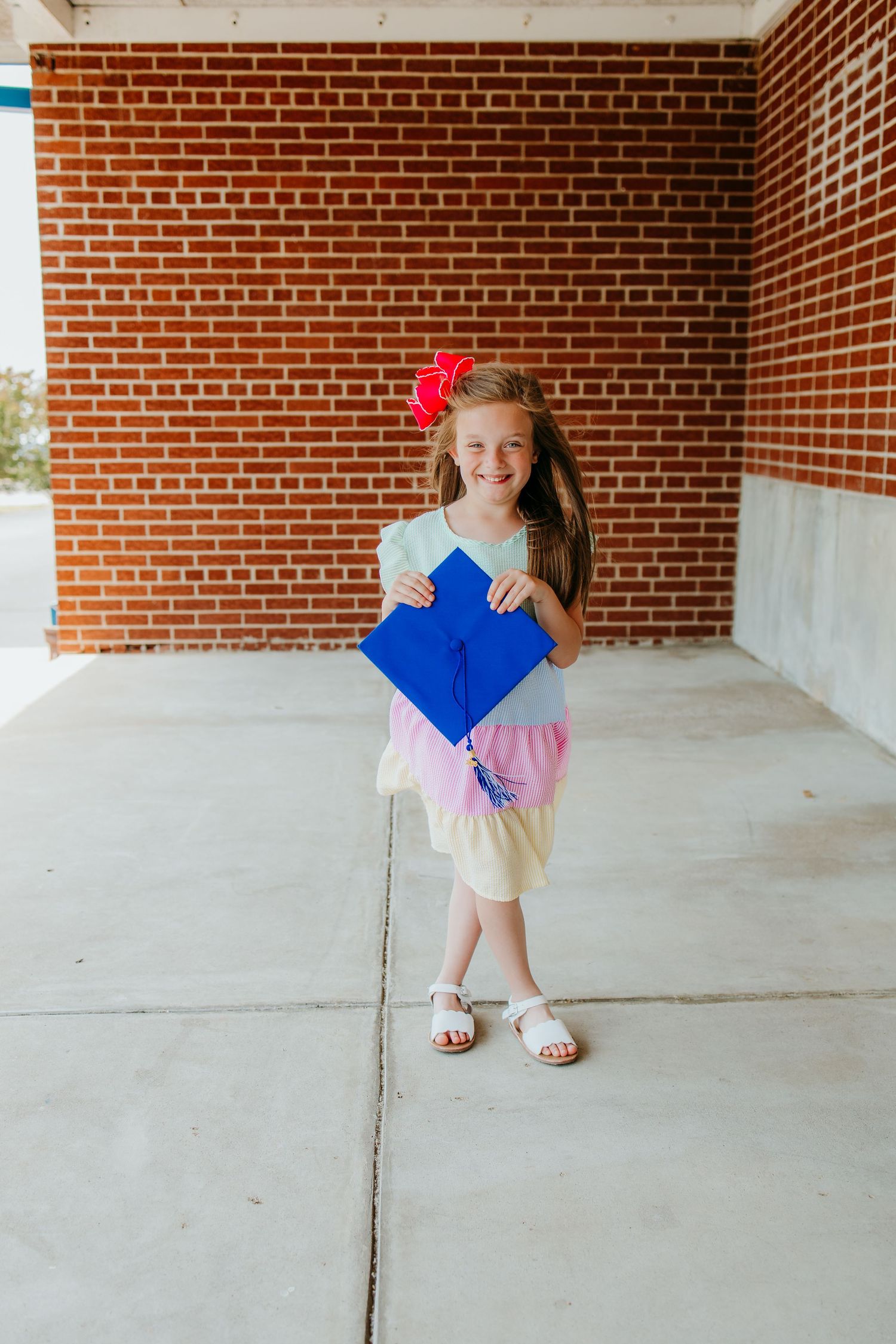 Young girl holding a blue graduation cap and tassel, smiling in front of a brick wall in a colorful dress.