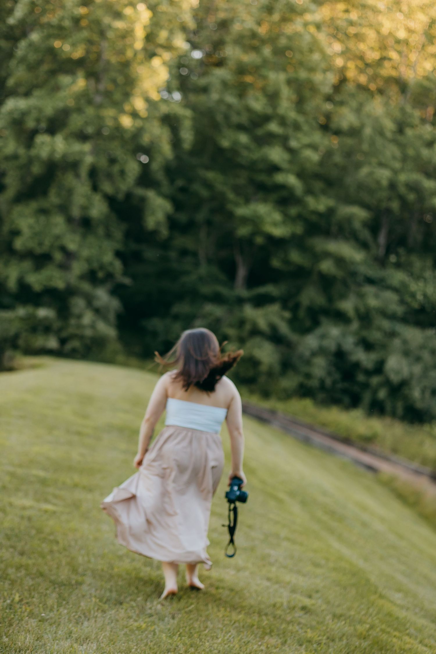 A woman in a flowy plaid skirt holds a camera while walking barefoot across a lush green lawn toward trees.
