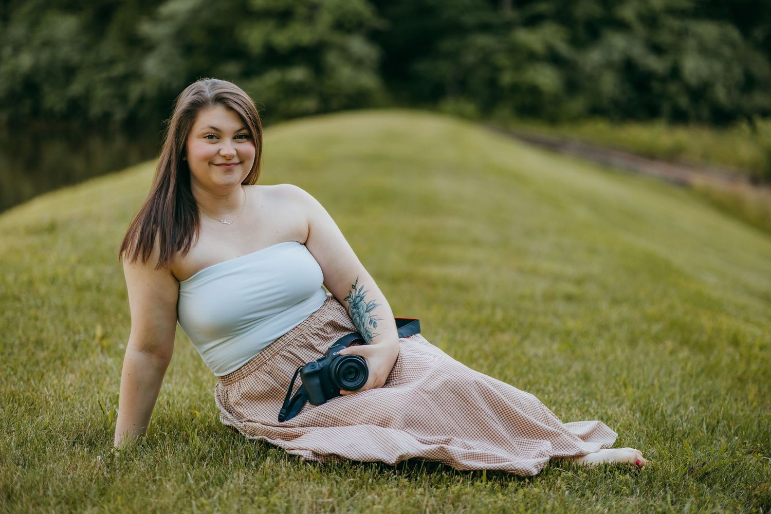 Woman in white tube top and plaid skirt sits on green grass hill holding a camera, smiling outdoors.