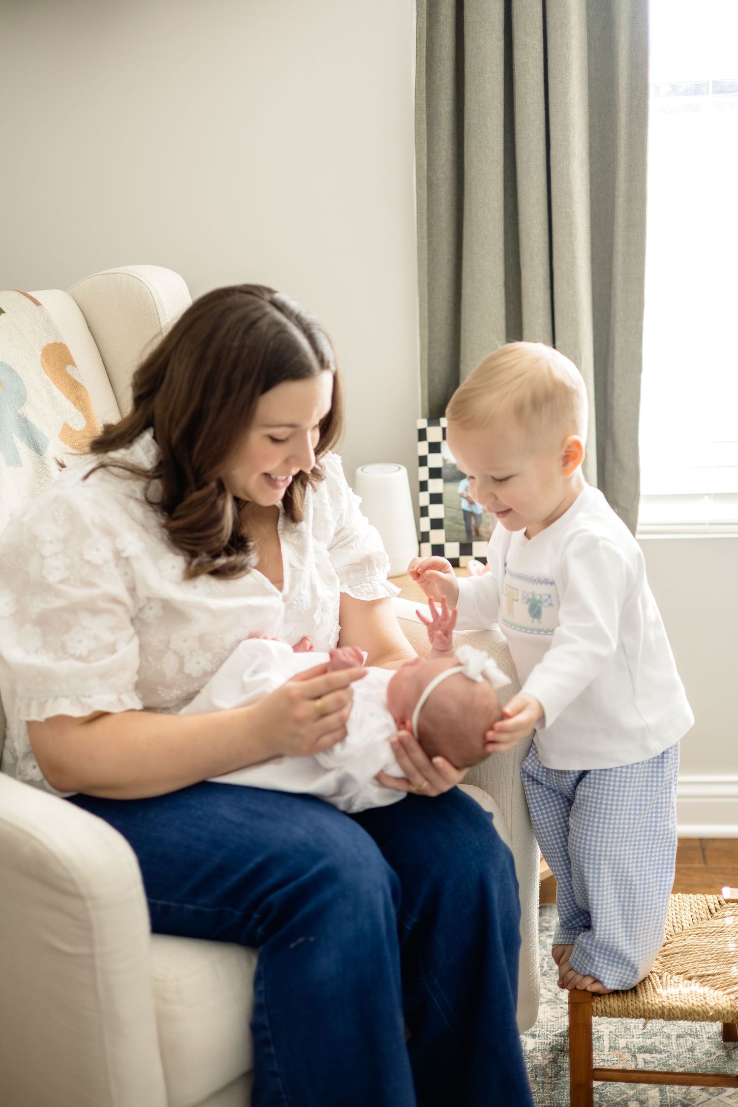 A mother sits in a nursery chair holding her newborn while a toddler boy smiles and touches the baby's hand.