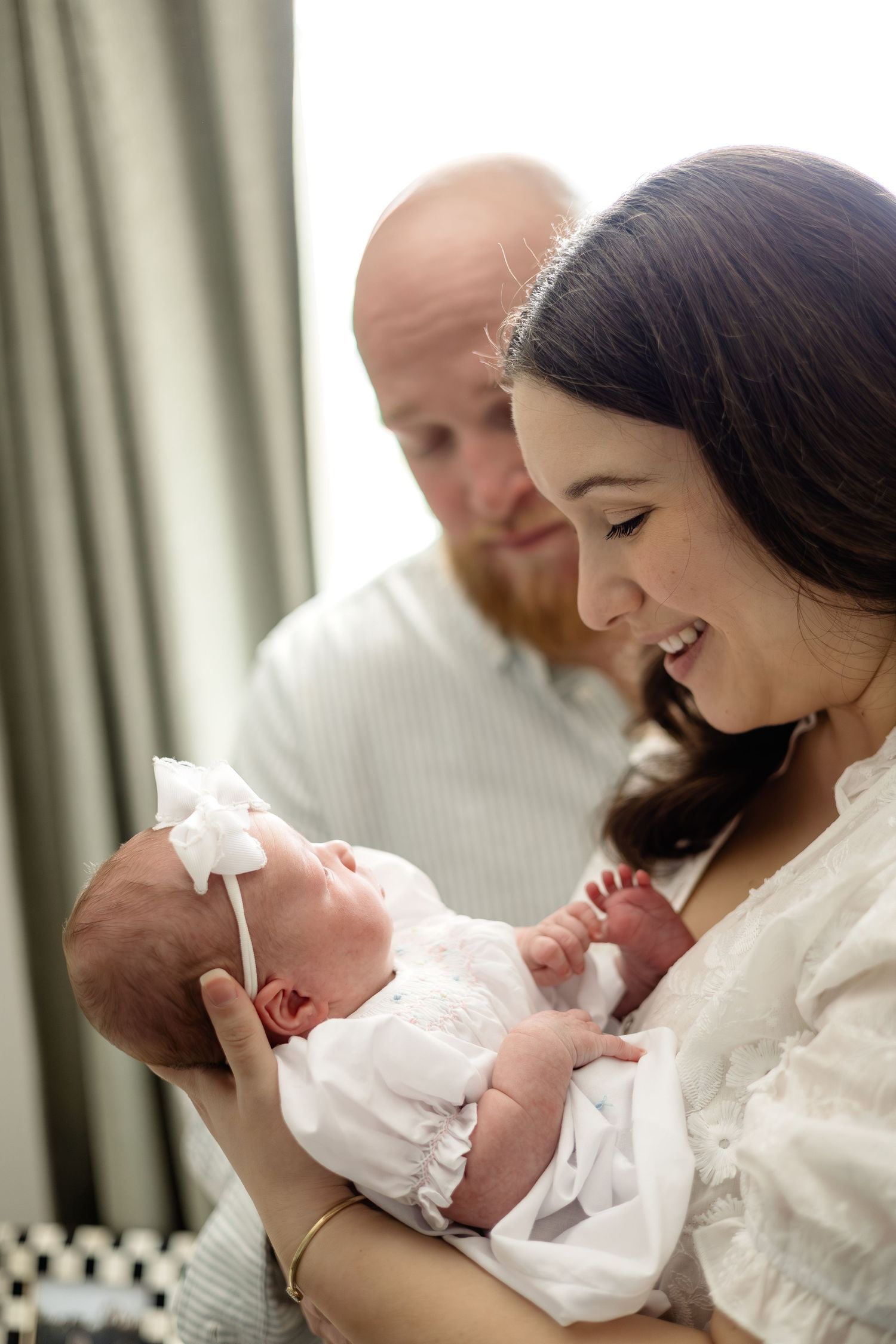 Parents smiling at their newborn baby girl dressed in white with a bow headband during a newborn photo session.