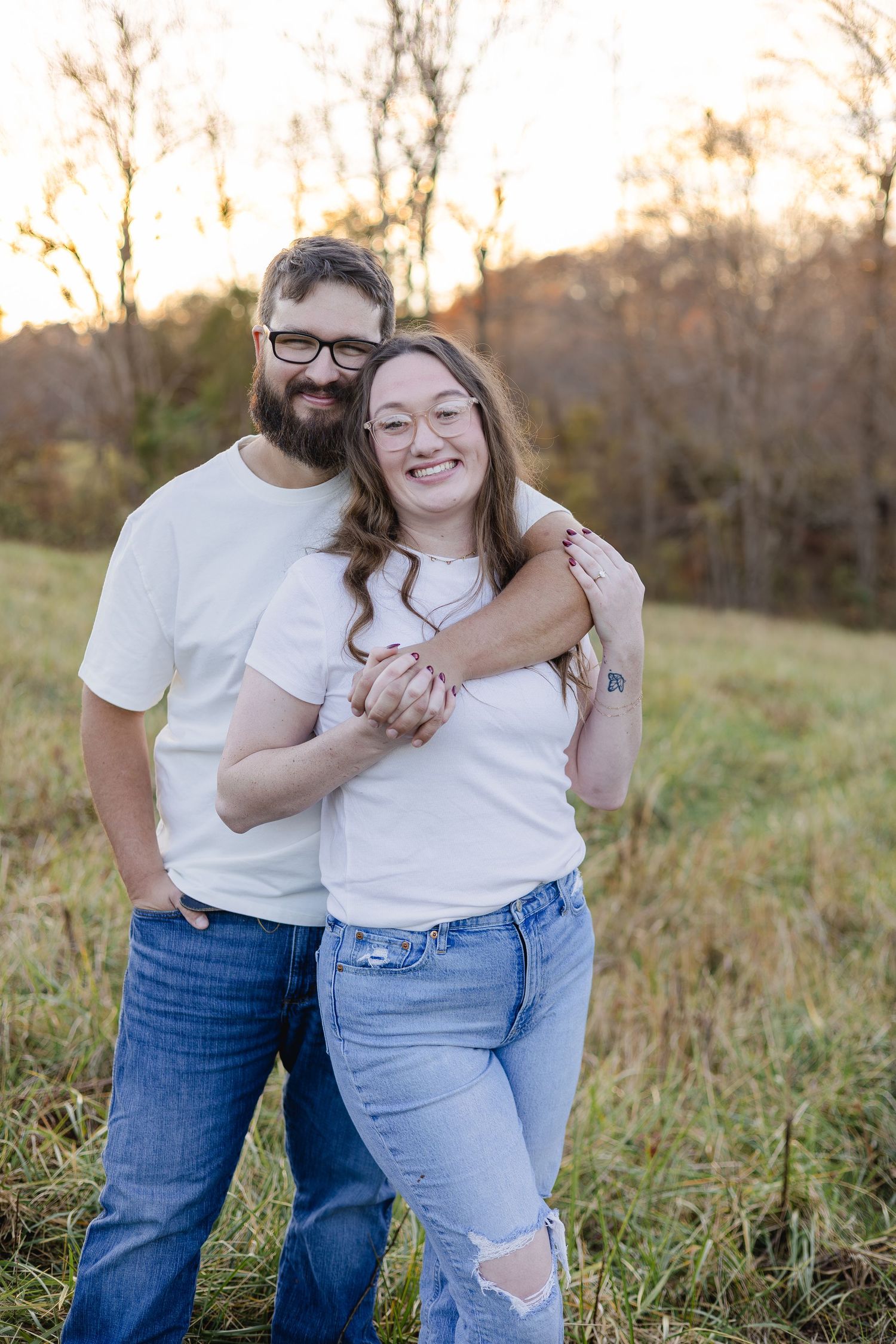 A smiling couple in matching white tees and jeans embrace outdoors in a golden autumn field.