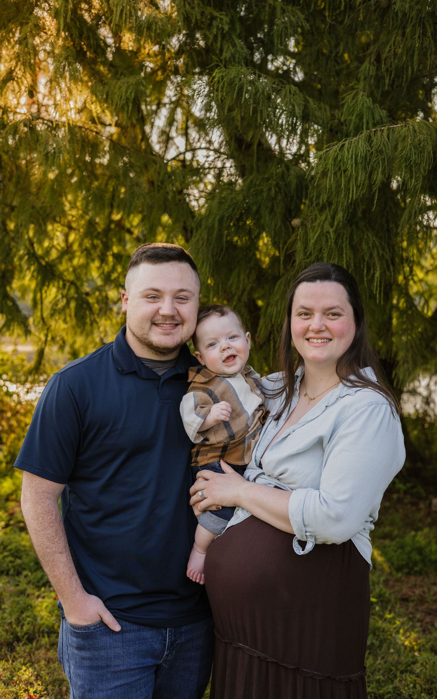 A smiling couple poses outdoors under a green tree, holding a happy baby, with the woman visibly pregnant.