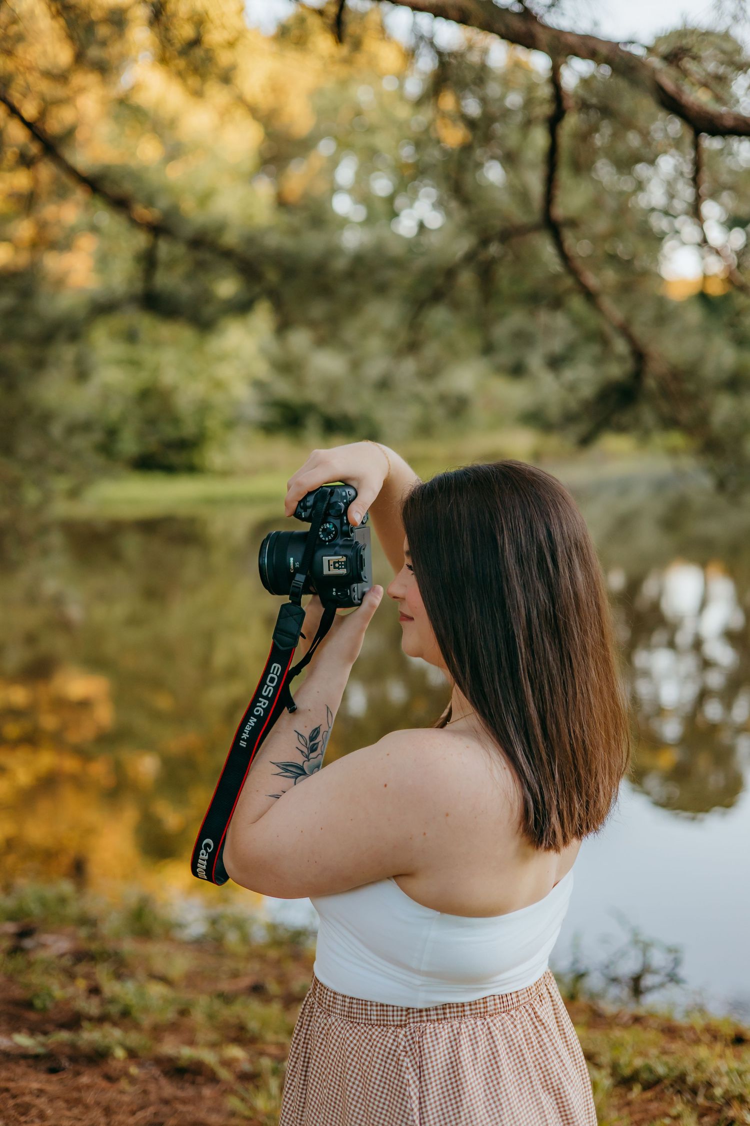 Woman with tattoo holding Canon DSLR camera outdoors near reflective pond surrounded by autumn trees.
