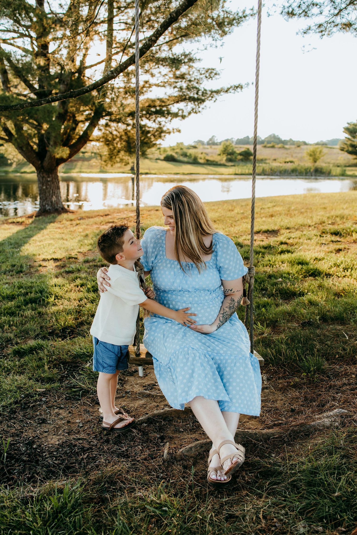 Pregnant woman in blue polka dot dress sitting on tree swing with young boy beside her near a serene lake.