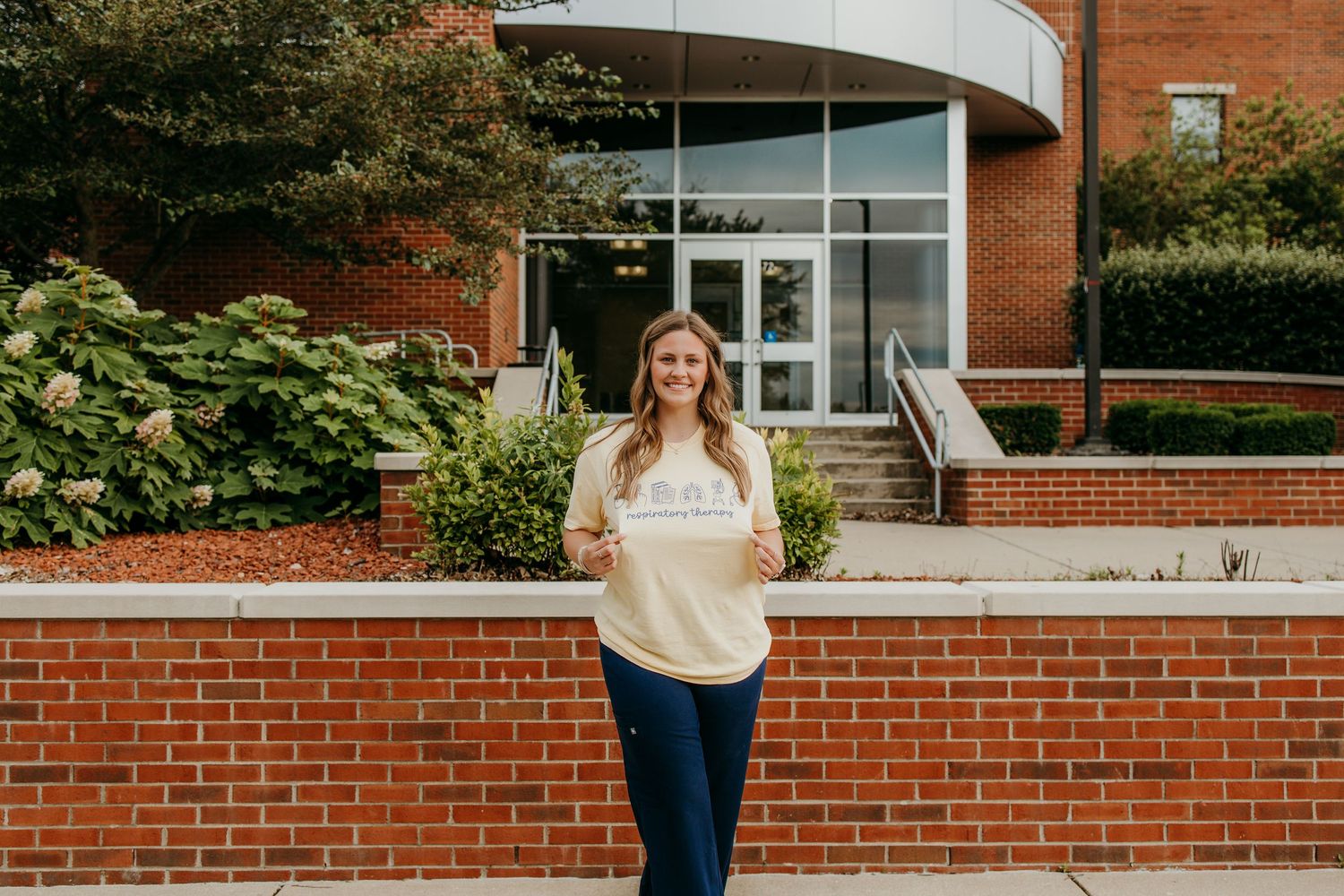 Young woman smiling in yellow shirt standing in front of brick building, college entrance outdoors.