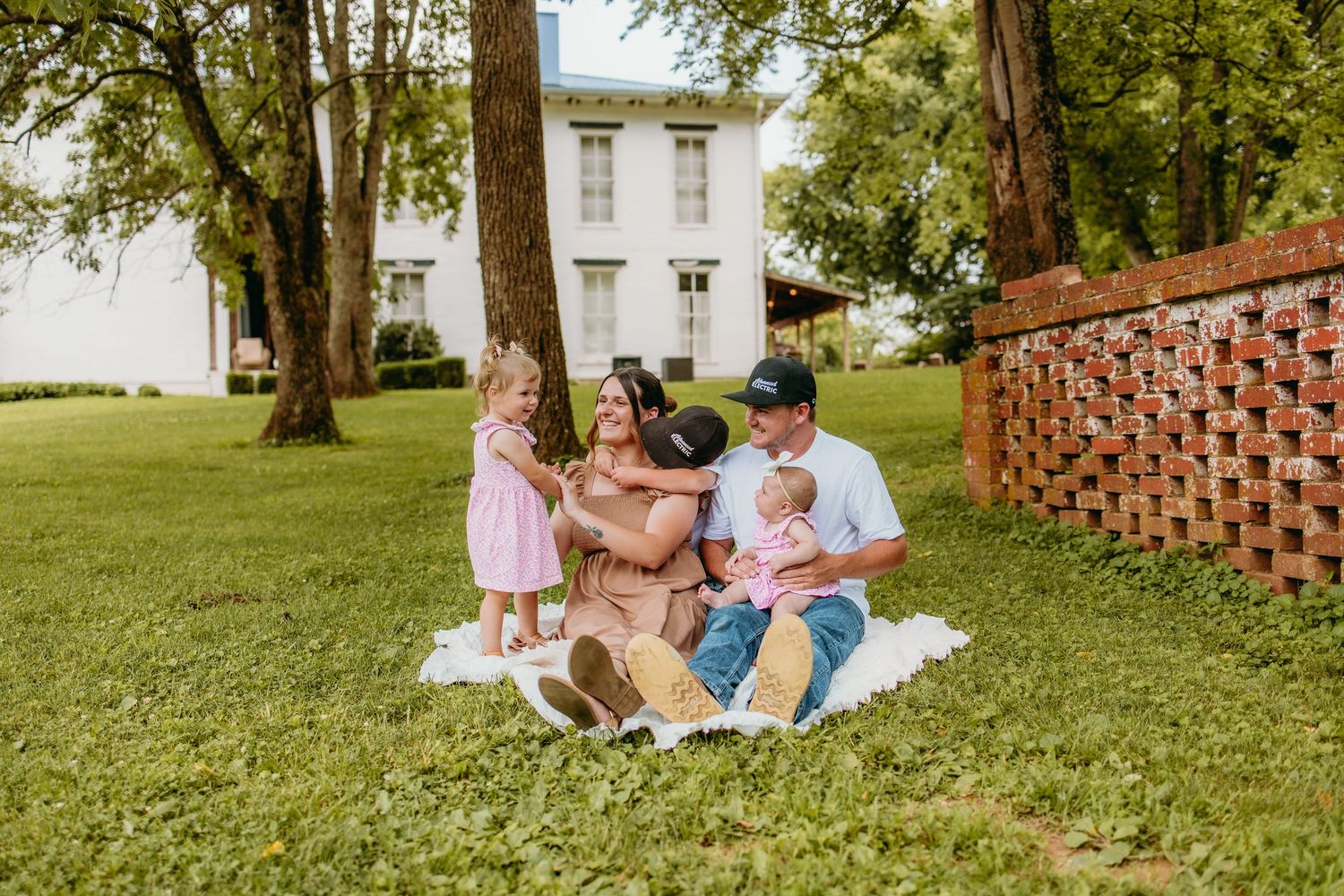 A happy family of five sits on a white blanket on green grass near a historic white building.