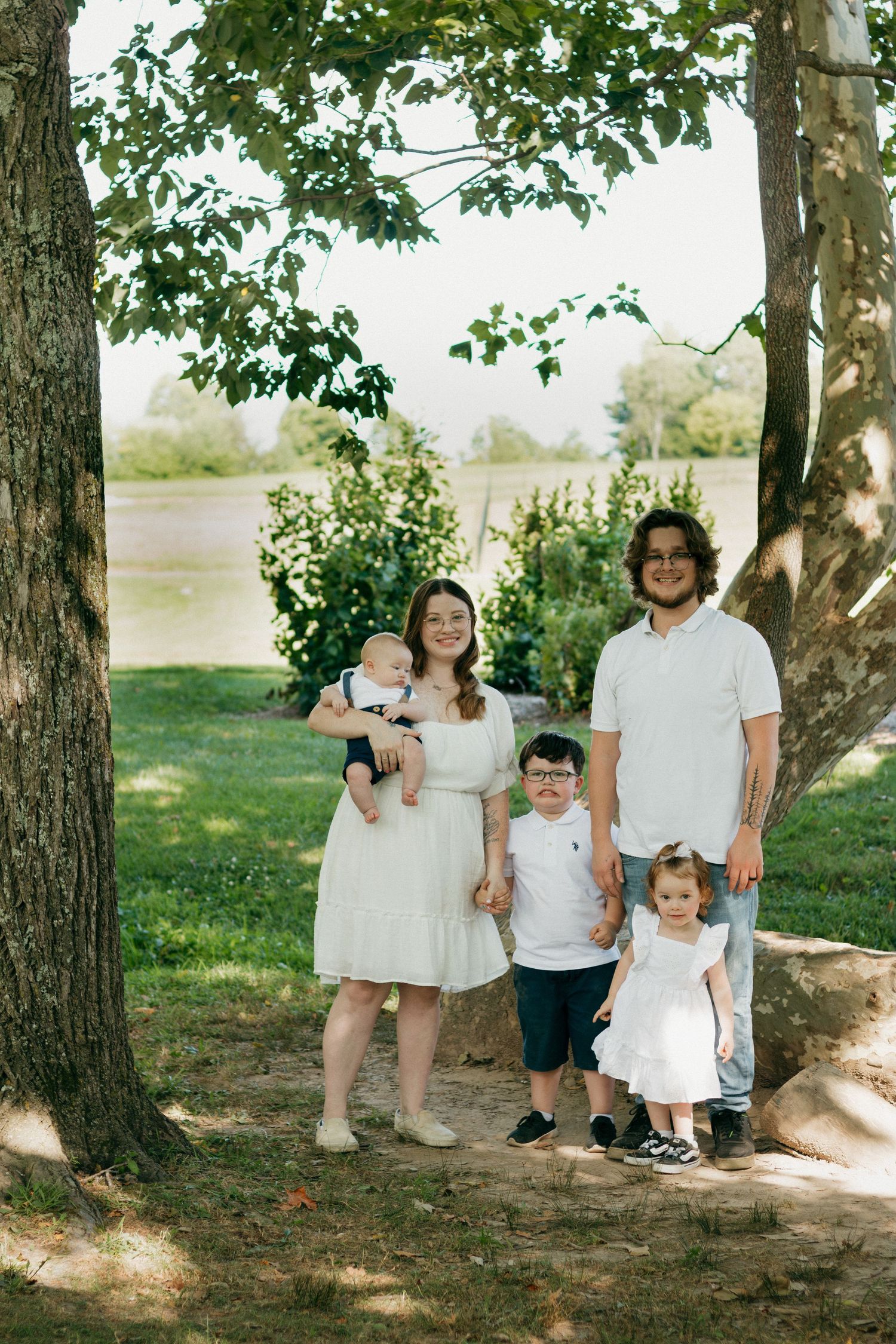 Family of five posing outdoors under trees in white outfits, parents holding young children in a lush green park setting.