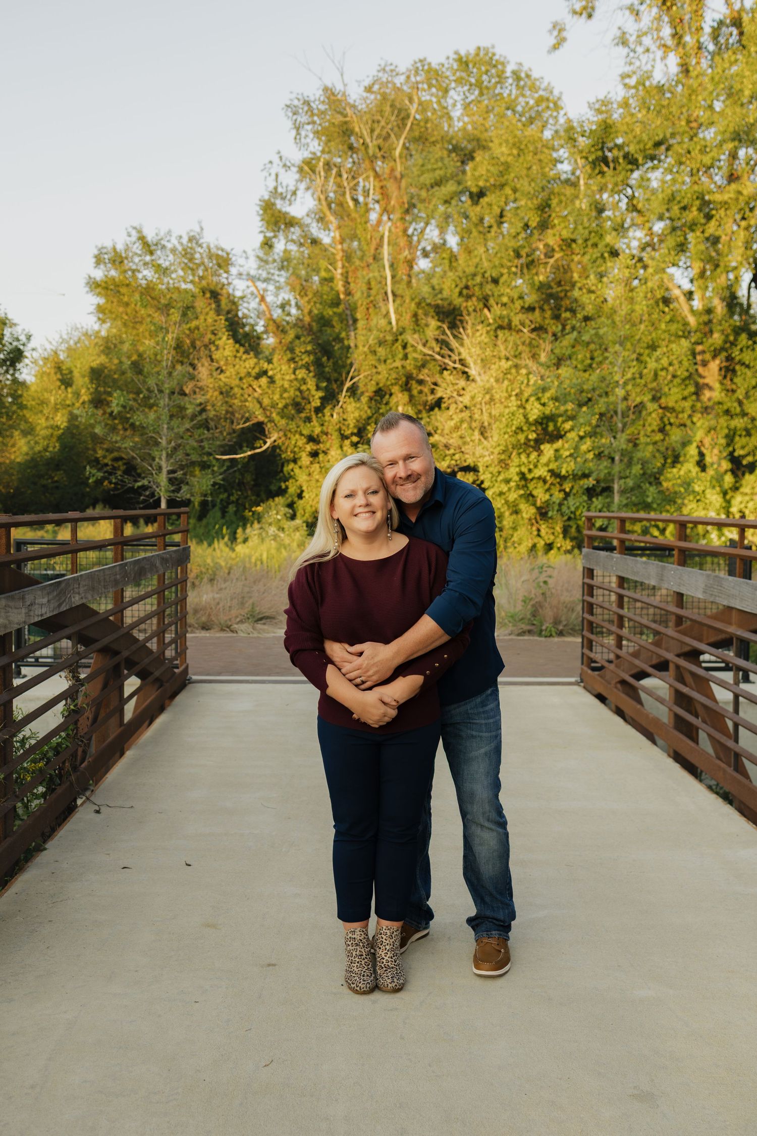 Couple embracing on a wooden bridge surrounded by lush green trees during golden hour outdoor photo session.