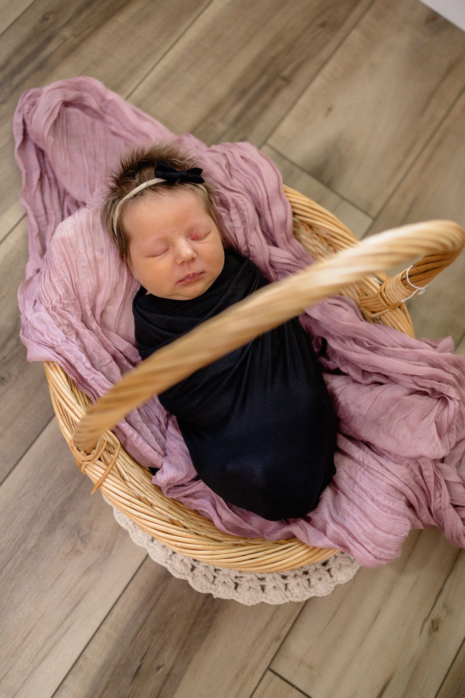 Sleeping newborn baby swaddled in black wrap, resting in wicker basket lined with pink cloth, wearing floral headband.