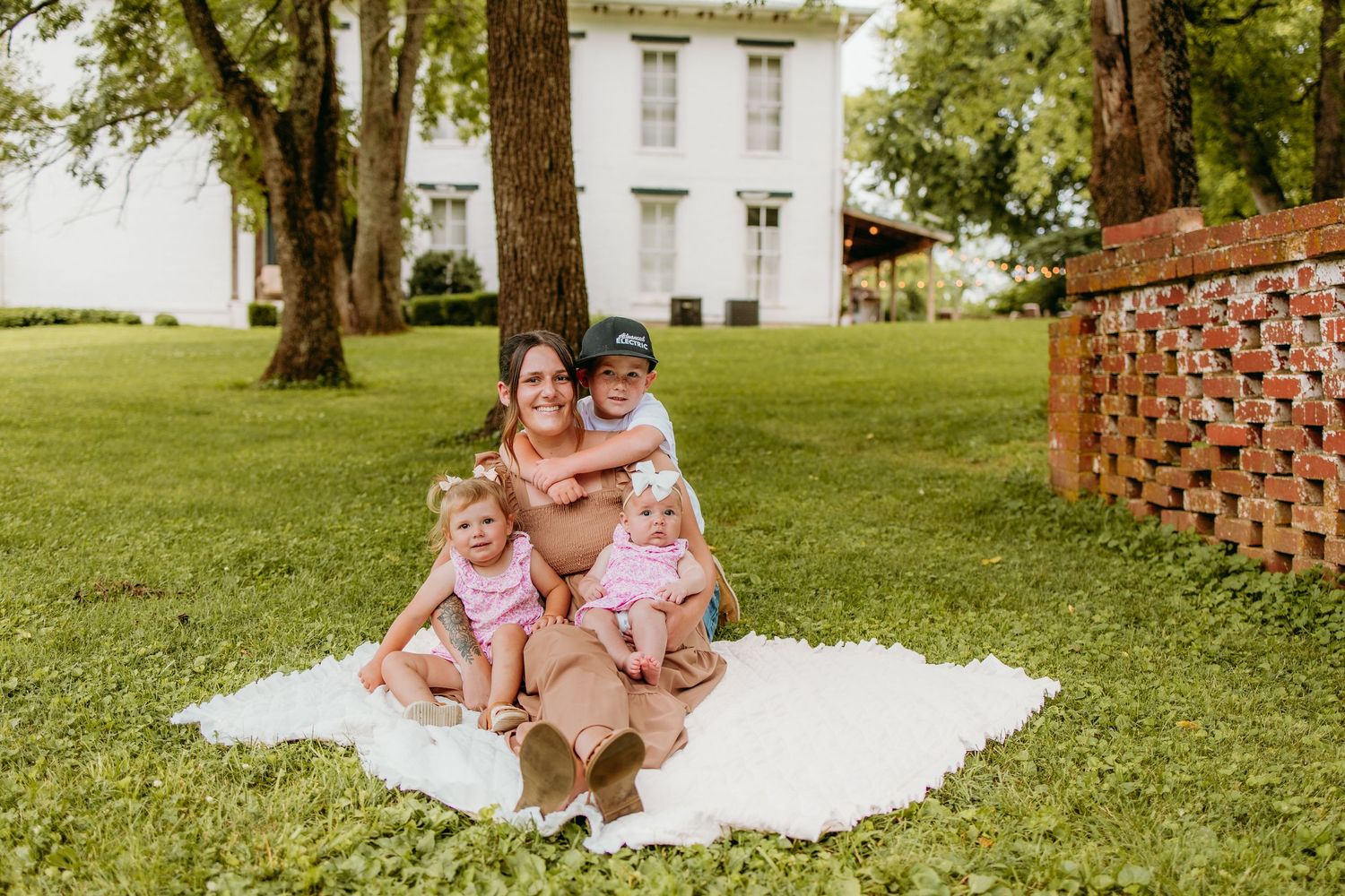 A mother sits on a white blanket outdoors with three young children in a lush green yard near a white house.