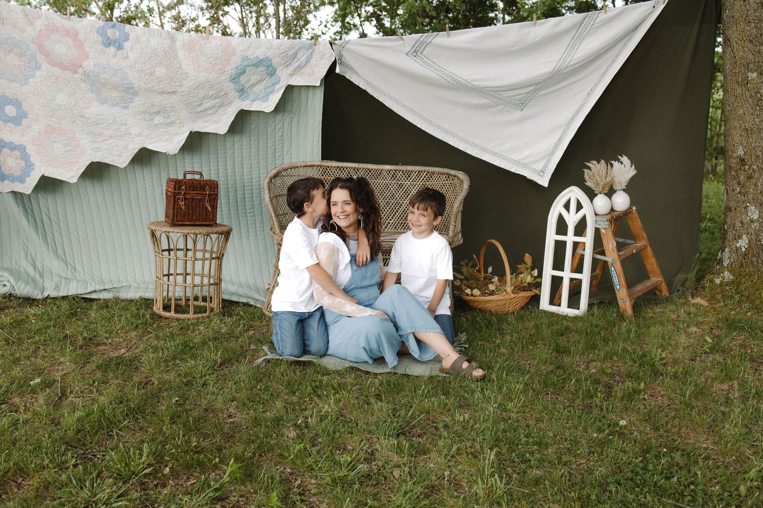 Family of three sitting on grass near a rustic tent with vintage decor, sharing a warm embrace outdoors.