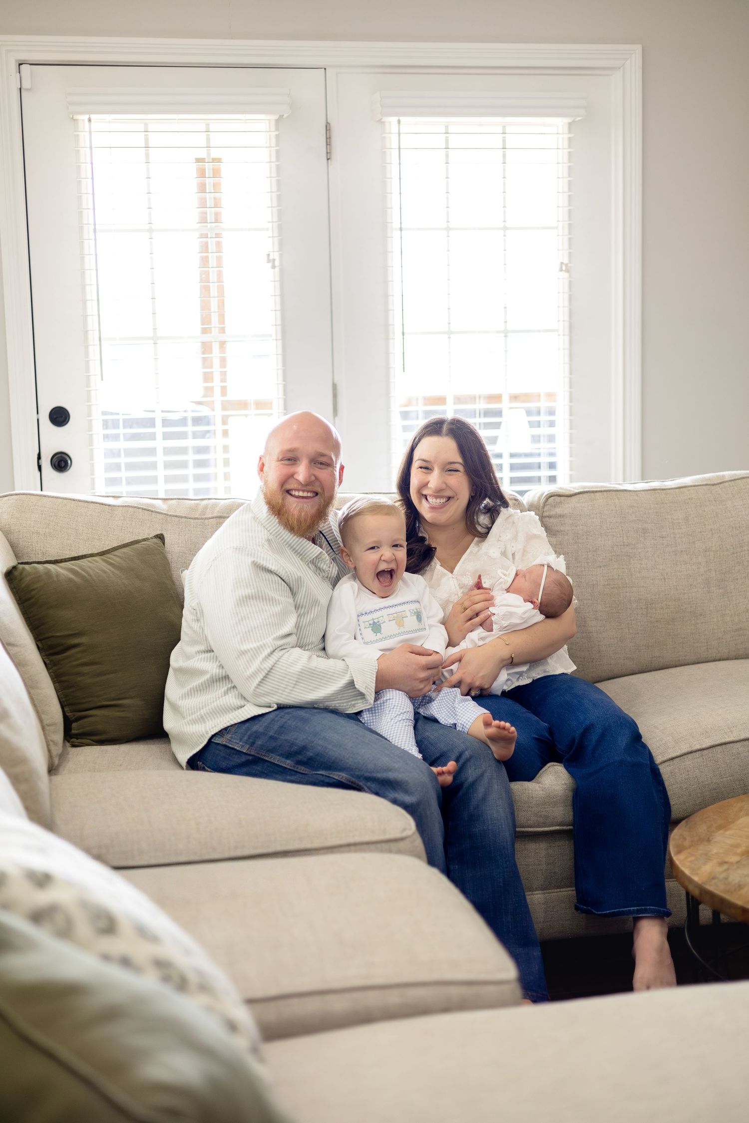 Happy family of four sitting on a beige sofa, parents smiling with a toddler and newborn baby in a bright living room.