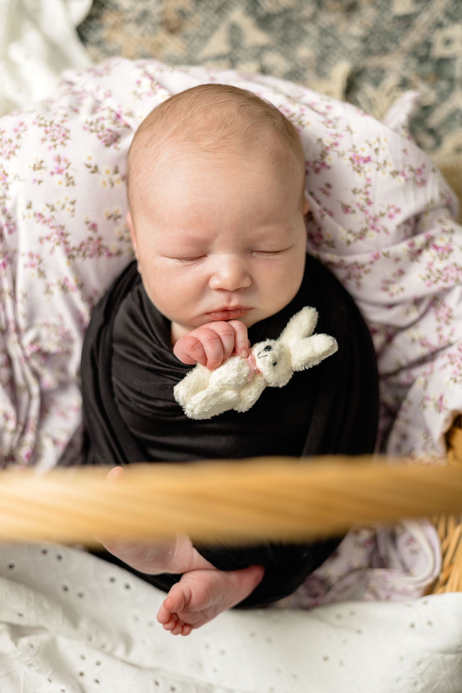 Sleeping newborn baby swaddled in black wrap holding a small white bunny toy, resting in a basket.