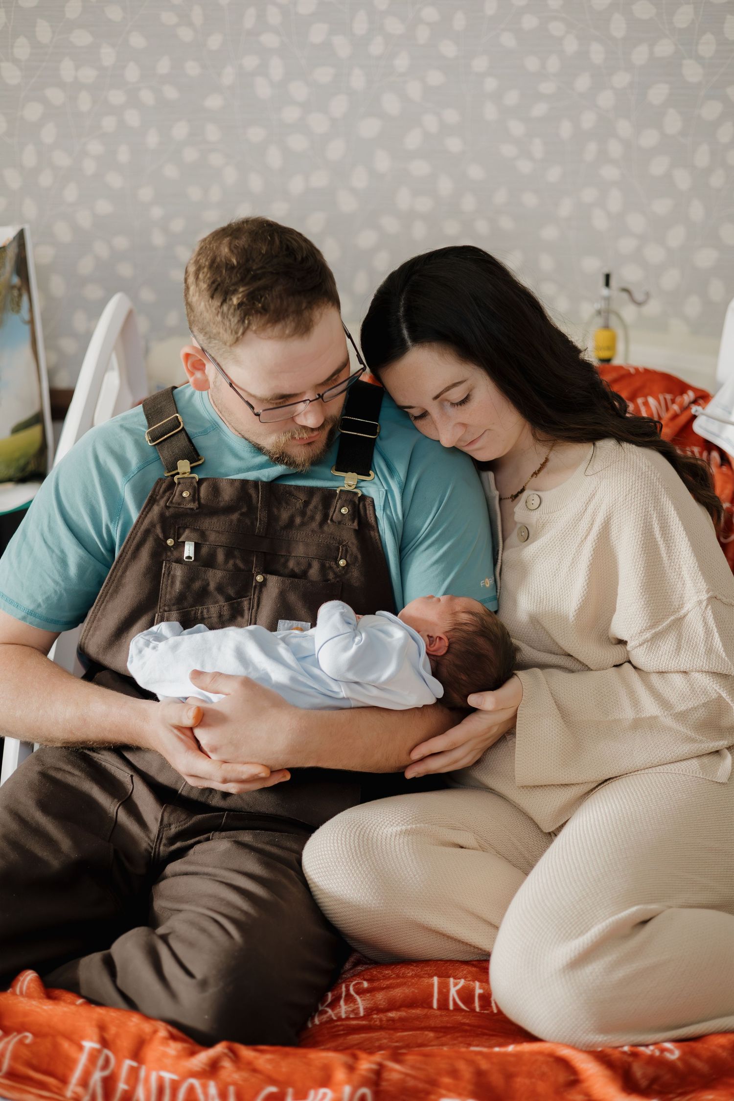 A couple sits together on a bed, lovingly gazing at their newborn baby wrapped in white.
