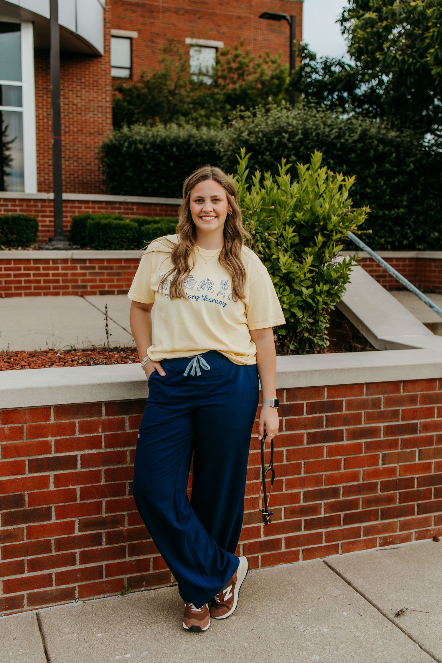 Young woman in yellow graphic tee and navy wide-leg pants leaning against brick wall outside building.
