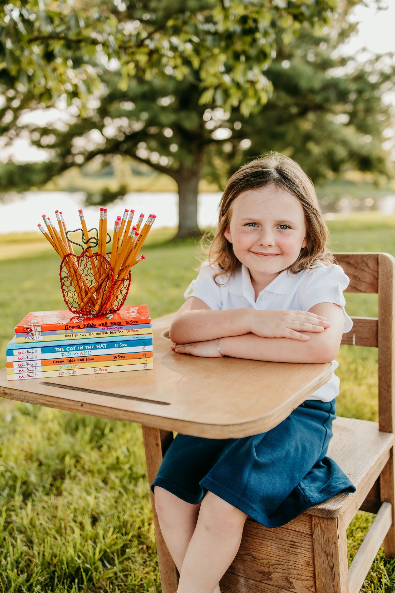 Young girl smiling at desk outdoors with Dr. Seuss books and pencils, back-to-school photo in park setting.