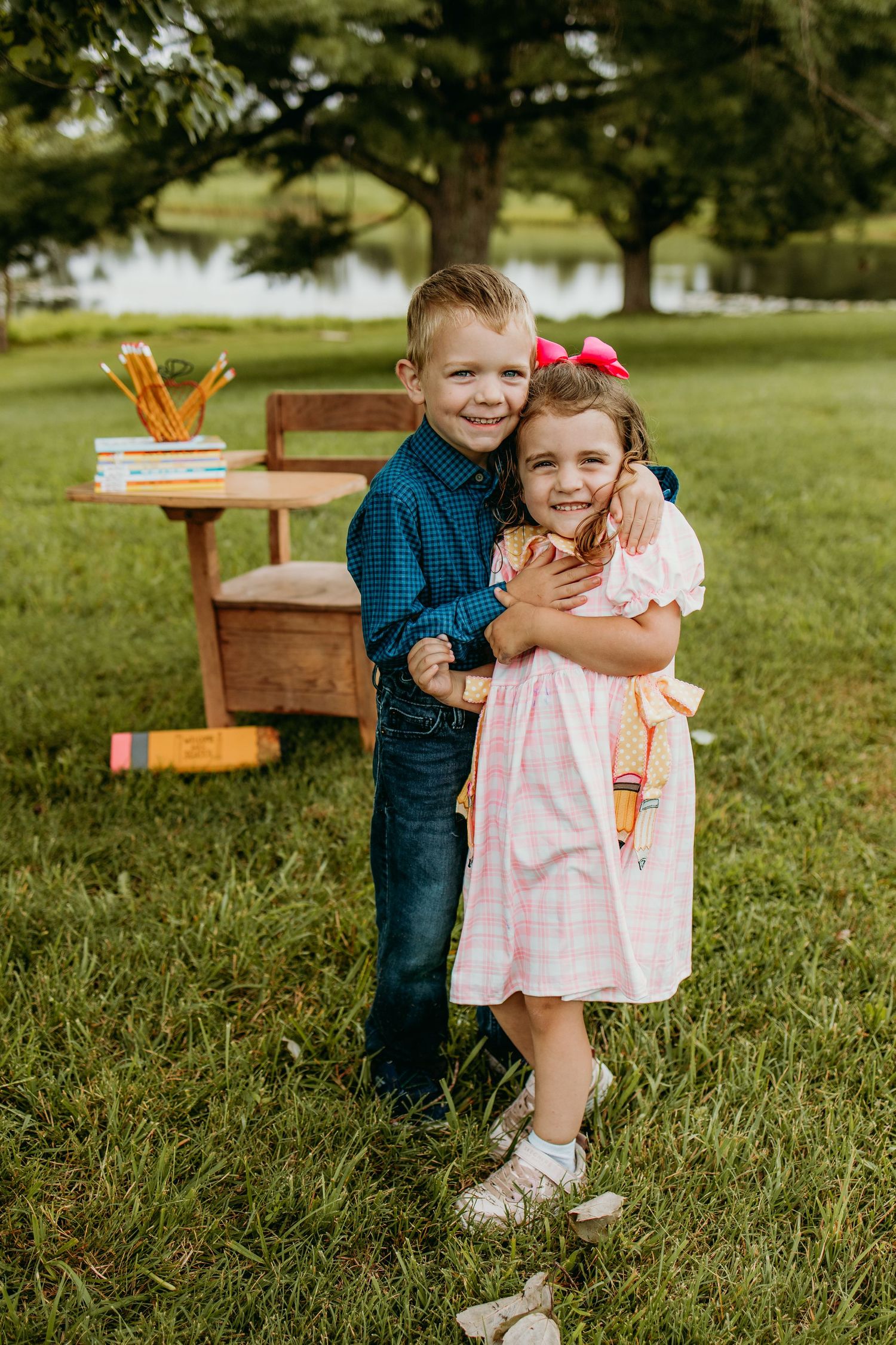 Two young children hugging outdoors near a wooden school desk with pencils, dressed in back-to-school outfits on green grass.