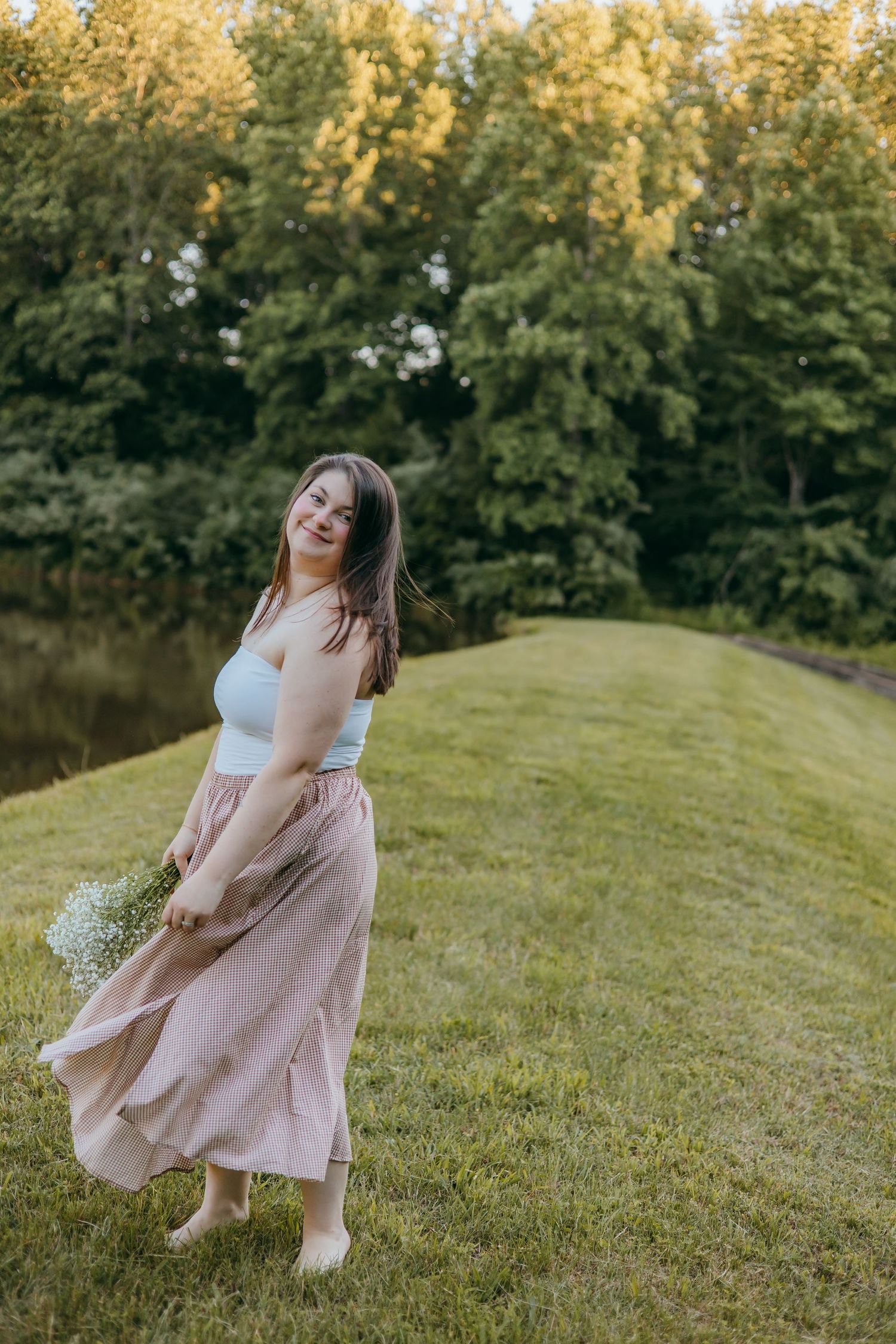 Young woman in plaid maxi skirt holding wildflowers, twirling barefoot on green grass with lush trees in background.