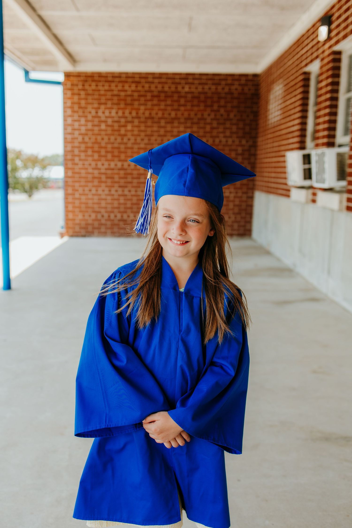Young girl smiling in blue graduation cap and gown standing in school hallway with brick wall background.