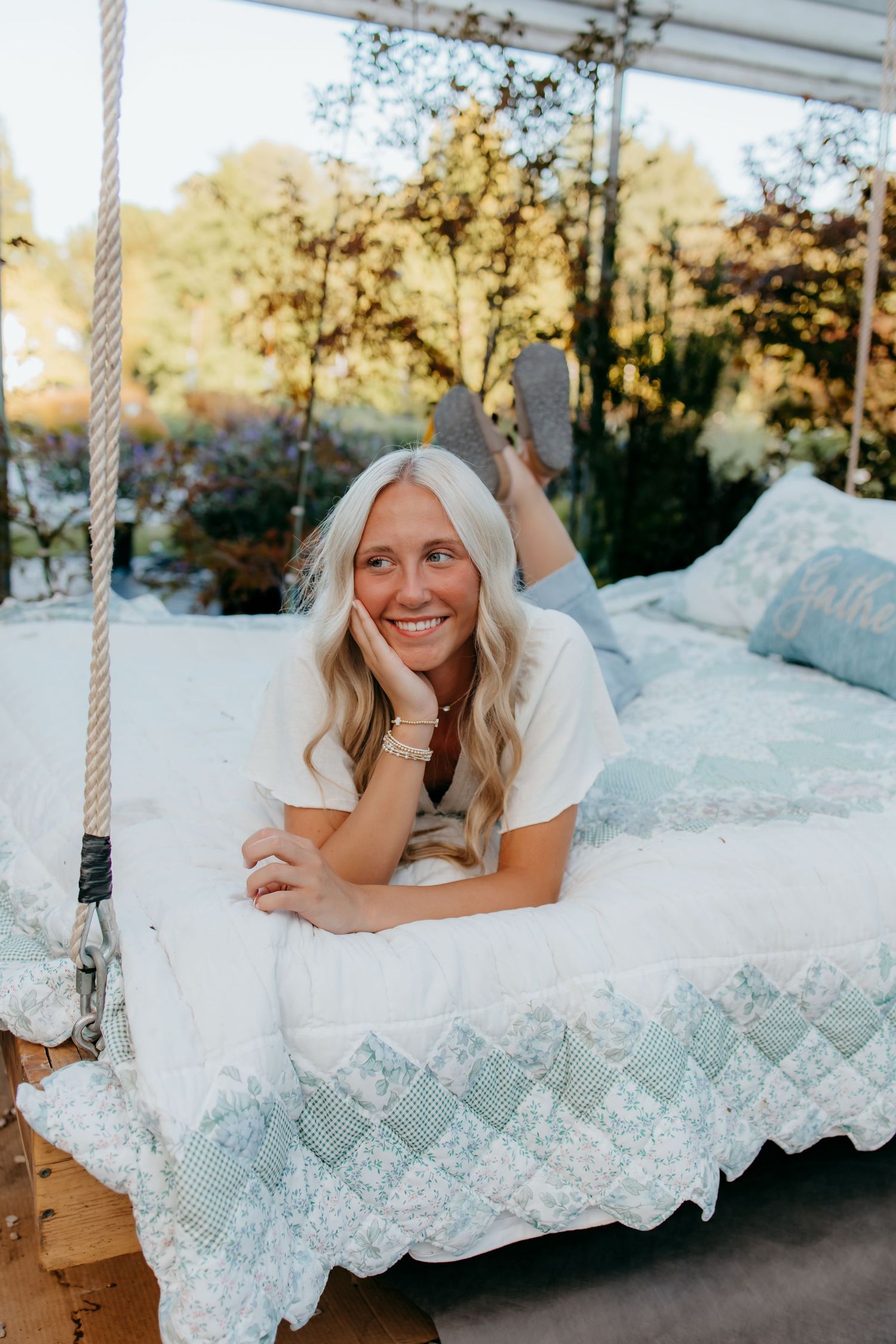 Smiling senior lying on a swing bed with blue and white quilt in a lush outdoor garden setting.