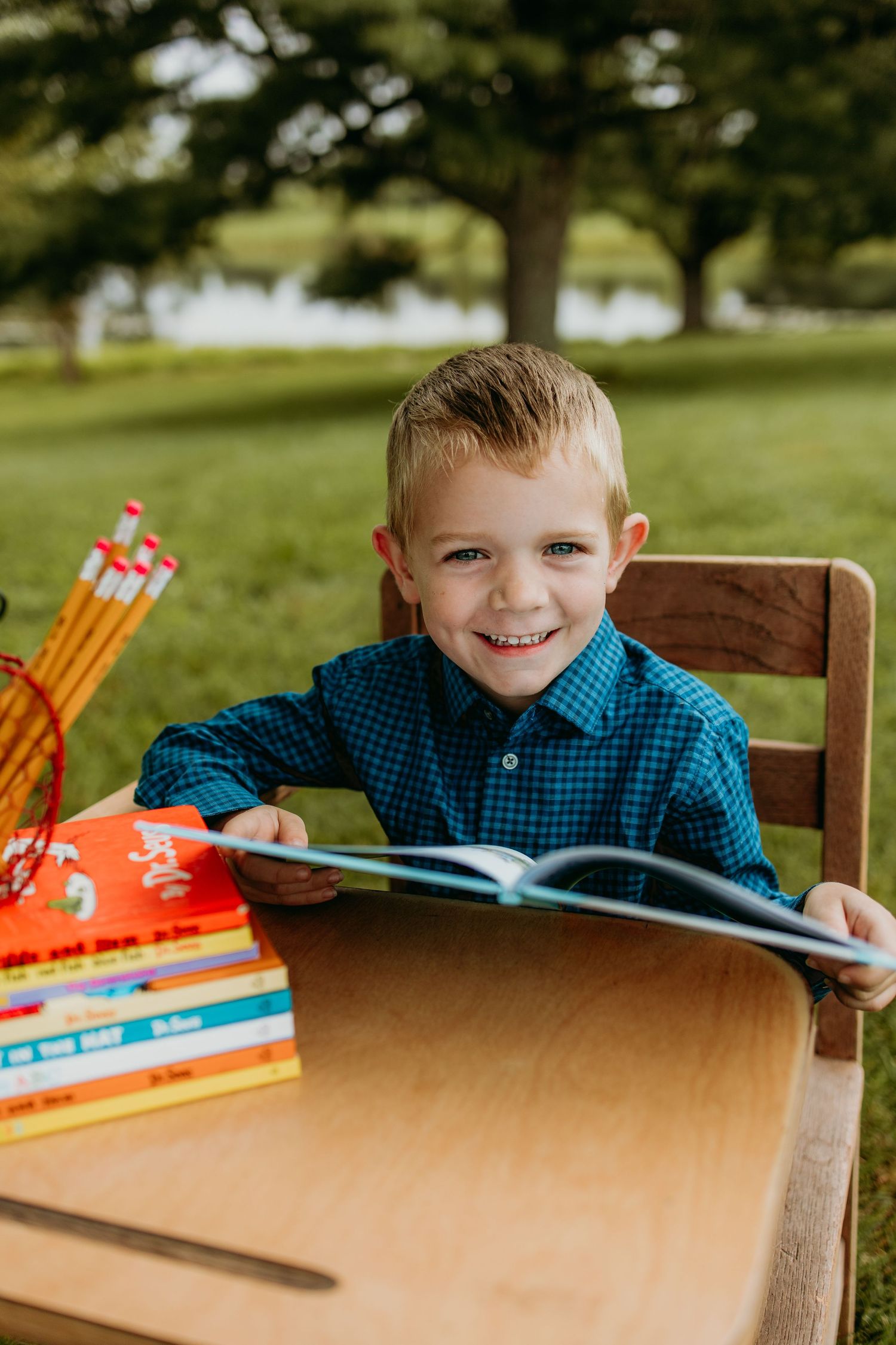 Smiling young boy in blue plaid shirt reading a book at a wooden desk outdoors with Dr. Seuss books and pencils.