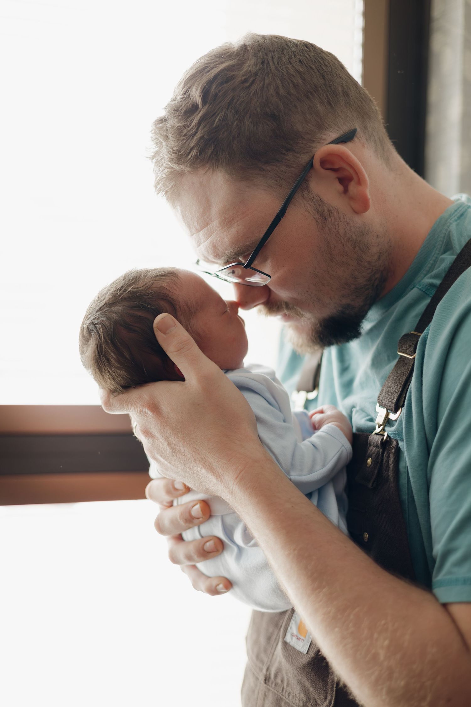 A bearded father with glasses tenderly kisses his newborn baby, cradling the infant gently in his hands.