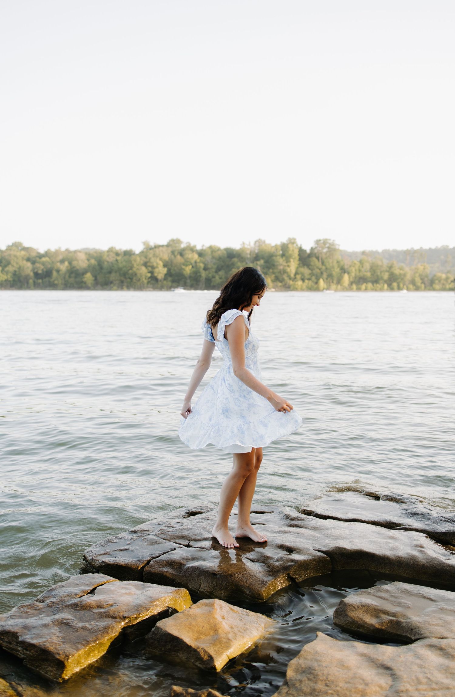 Young woman in white dress standing on rocks by a serene lake, surrounded by lush green trees.