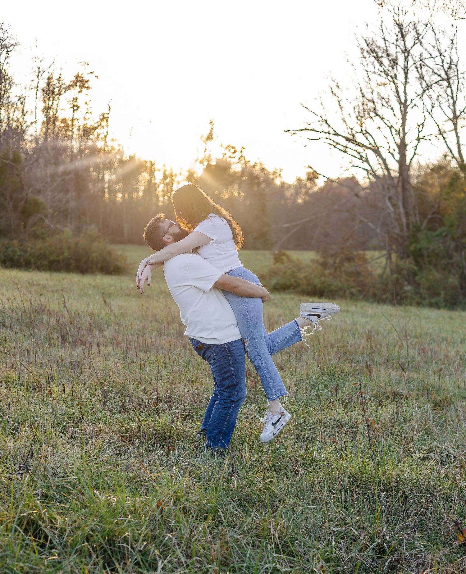 A couple shares a romantic embrace in a sunlit field, the man lifting the woman as golden hour light glows behind them.