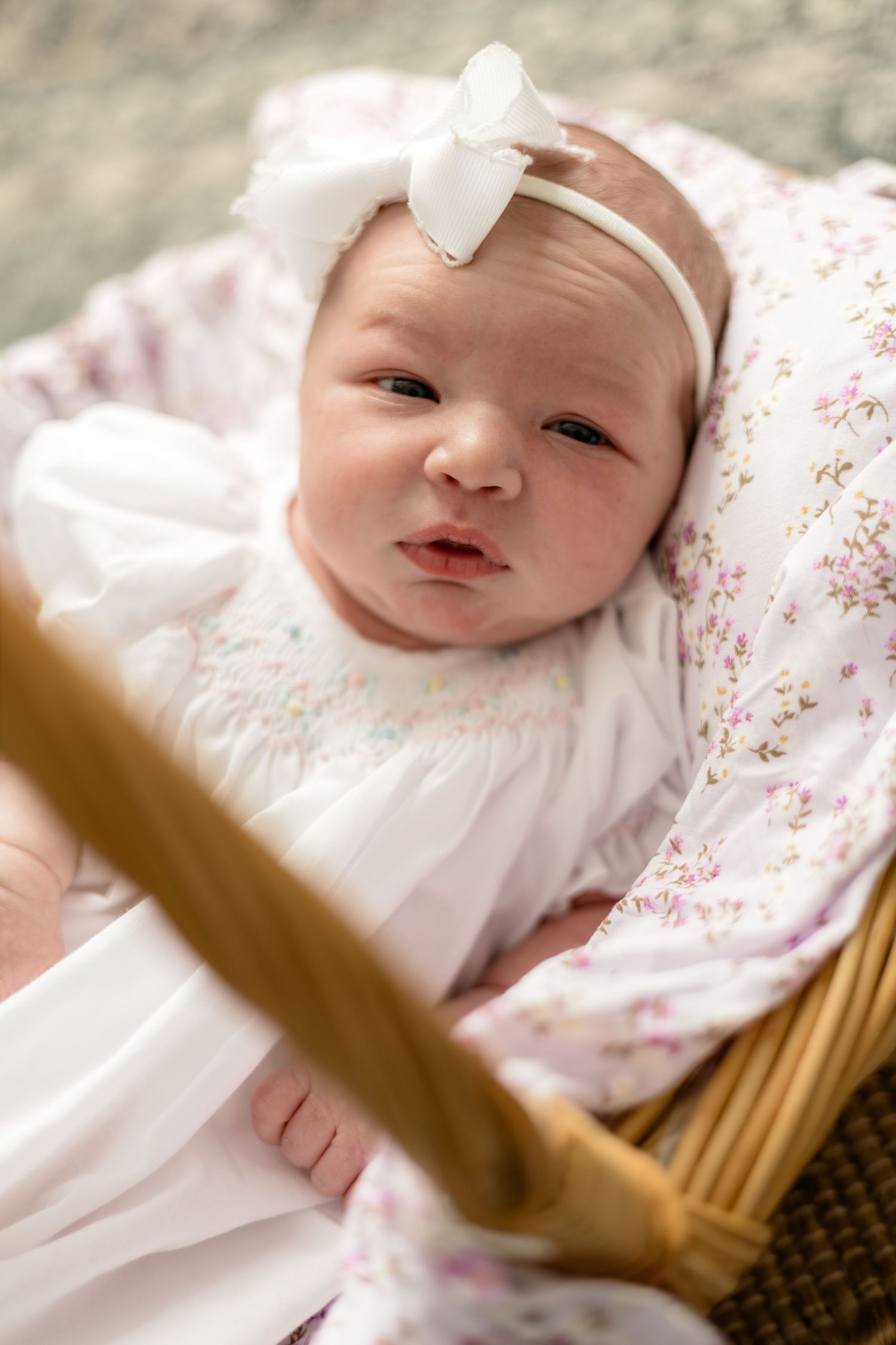 Newborn baby girl in white dress and bow headband resting in a wicker bassinet with floral blanket.