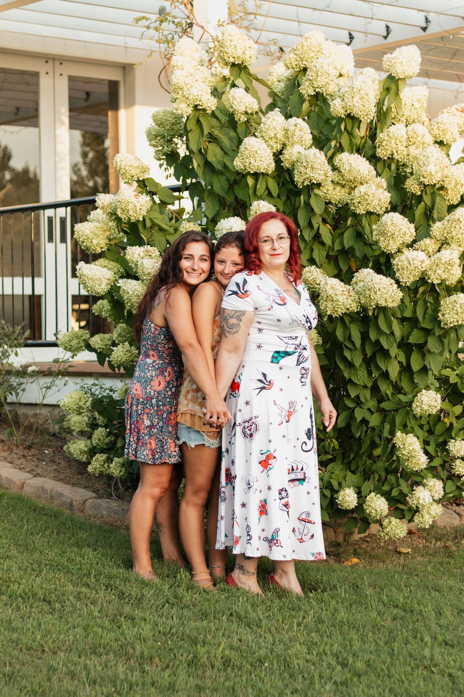 Three women pose together in front of a large blooming hydrangea bush outside a home.