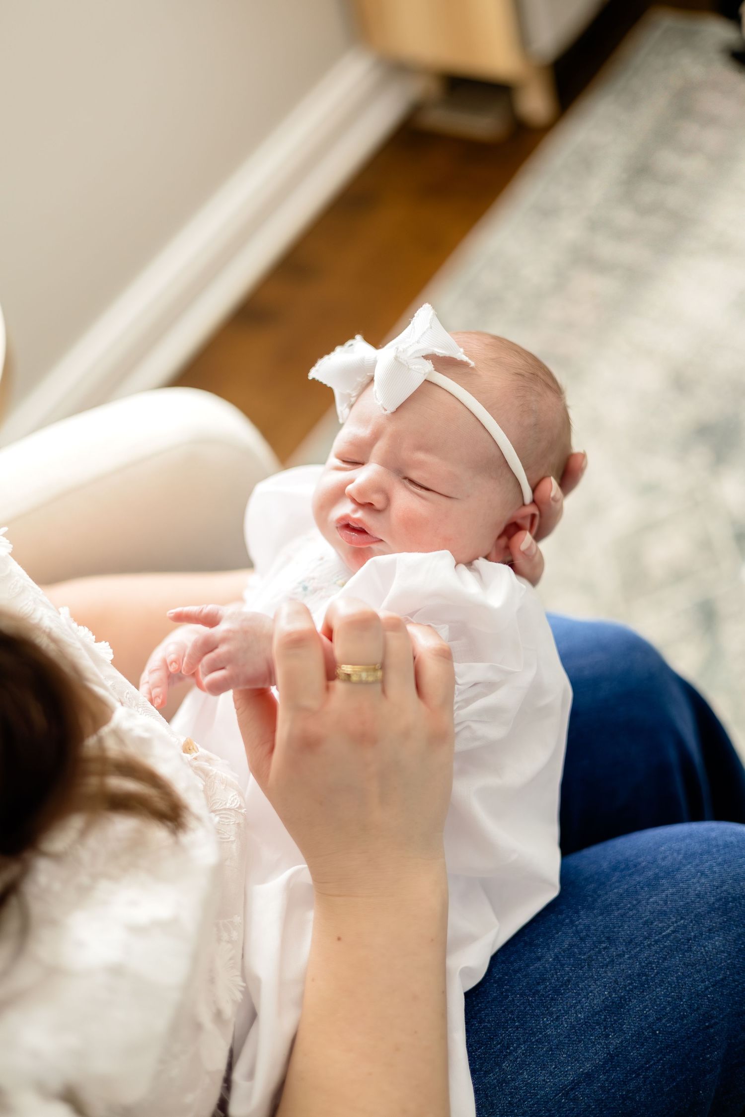 A newborn baby girl in a white dress and bow headband smiles while being cradled by her mother indoors.