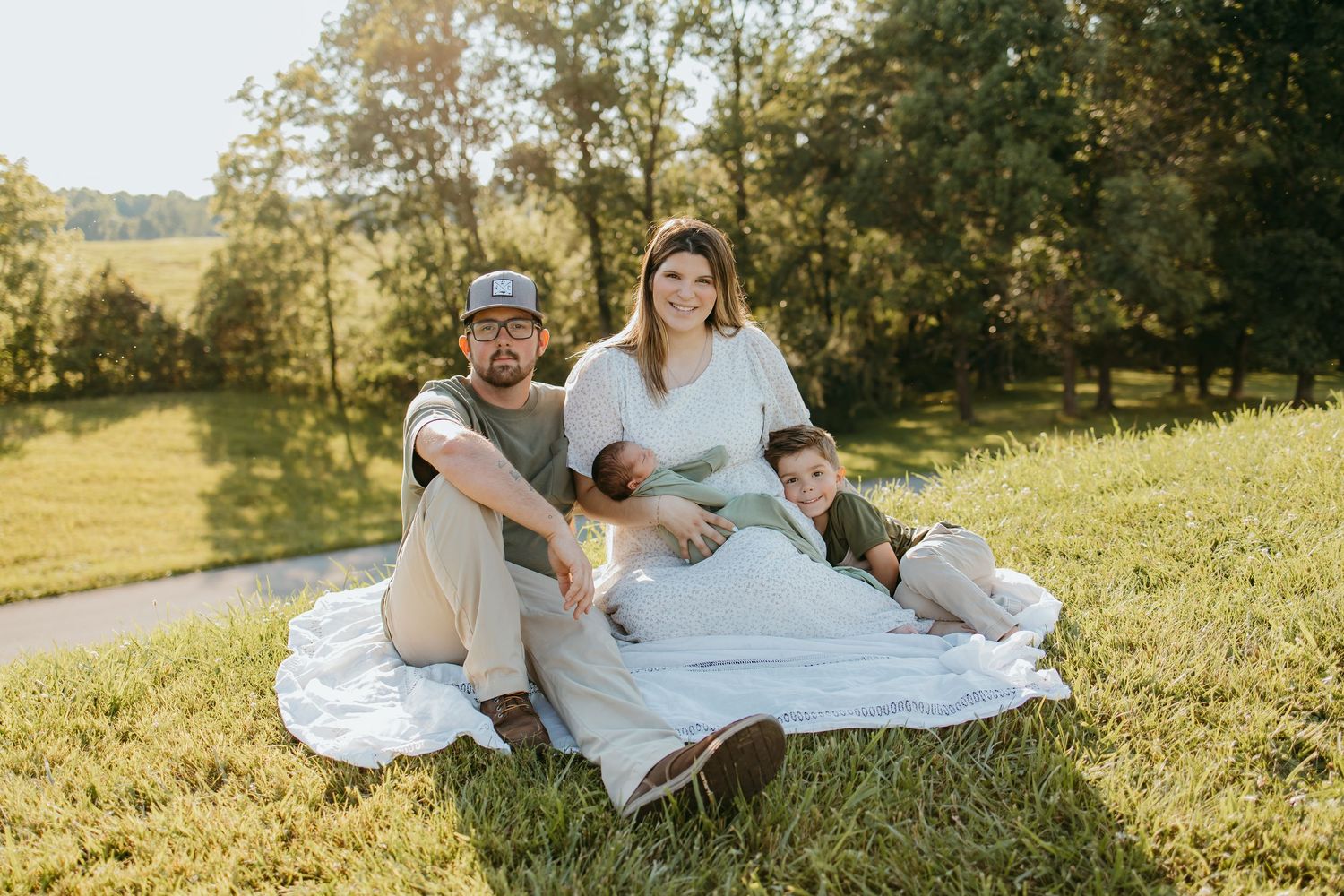 Family of four sitting on white blanket in sunny green field, parents holding newborn baby with young child beside them.