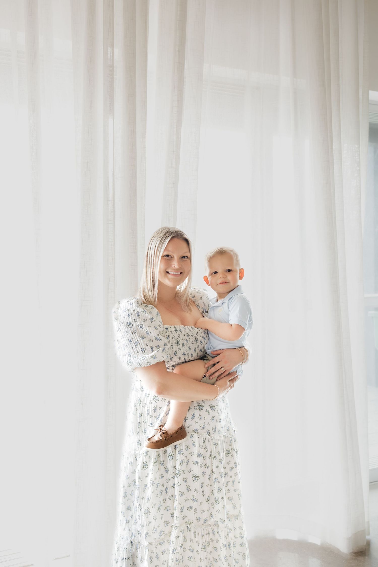 Smiling blonde mother in floral dress holding baby boy in blue outfit near sheer white curtains.