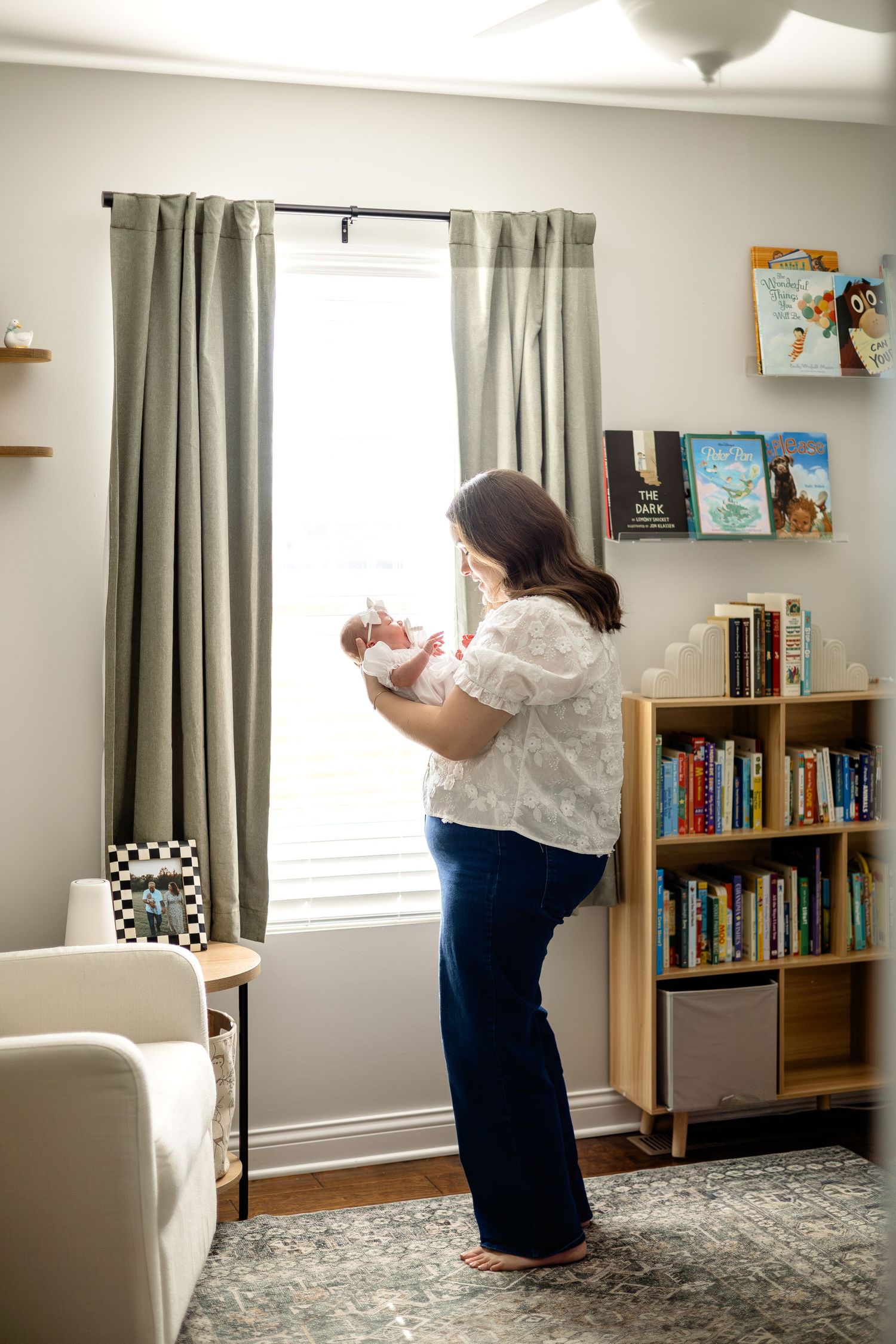 A mother stands by a window holding her newborn baby in a cozy nursery with bookshelves and sage green curtains.