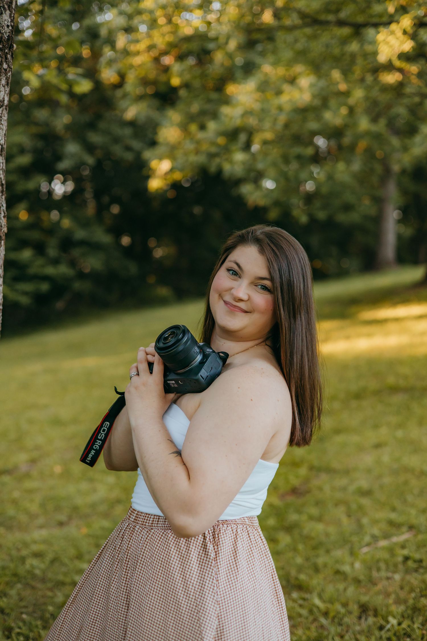 Young woman in white top and plaid skirt smiling while holding a DSLR camera outdoors in a green park.