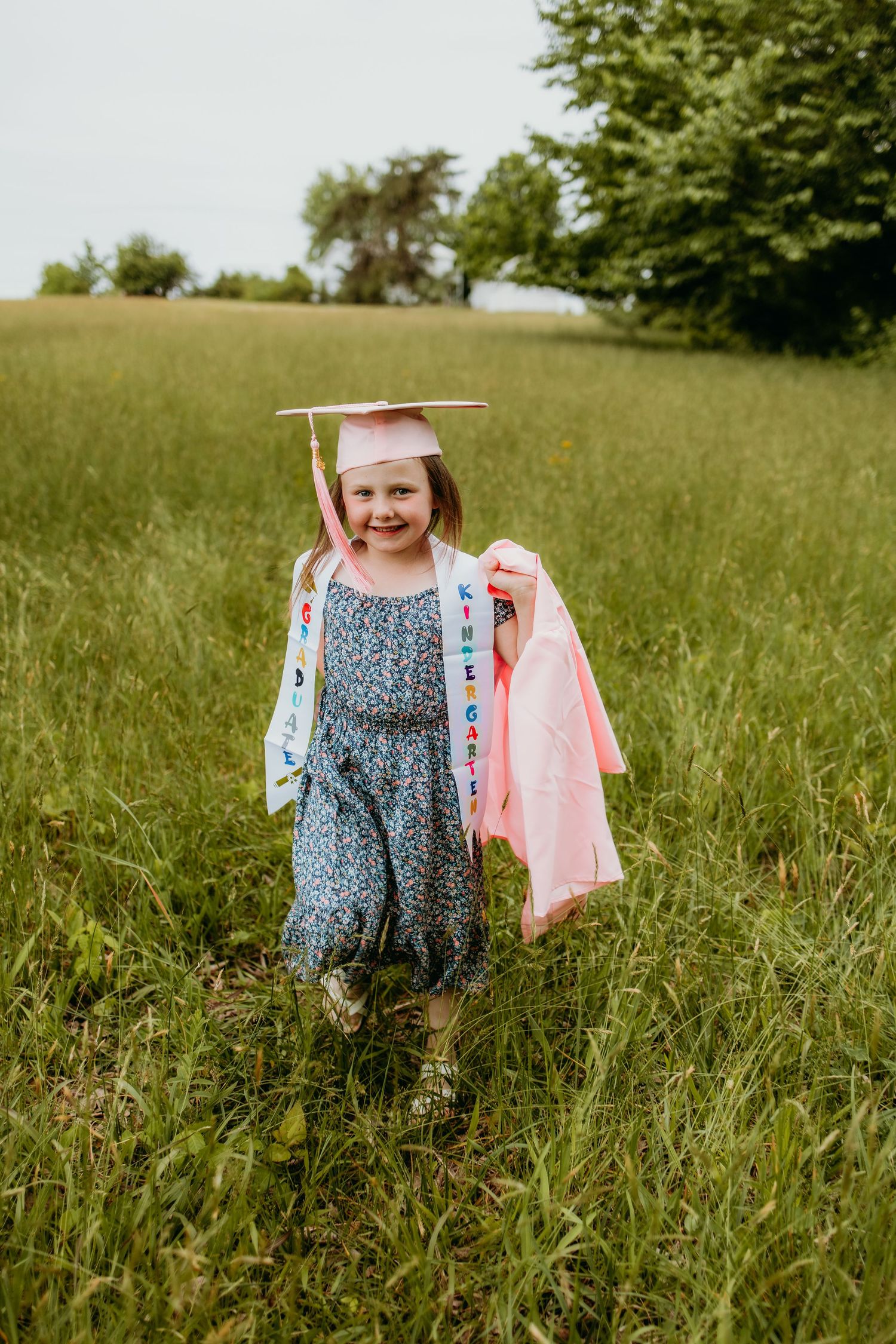 Young girl in graduation cap and sash smiles proudly in green field, holding pink gown on cloudy day.