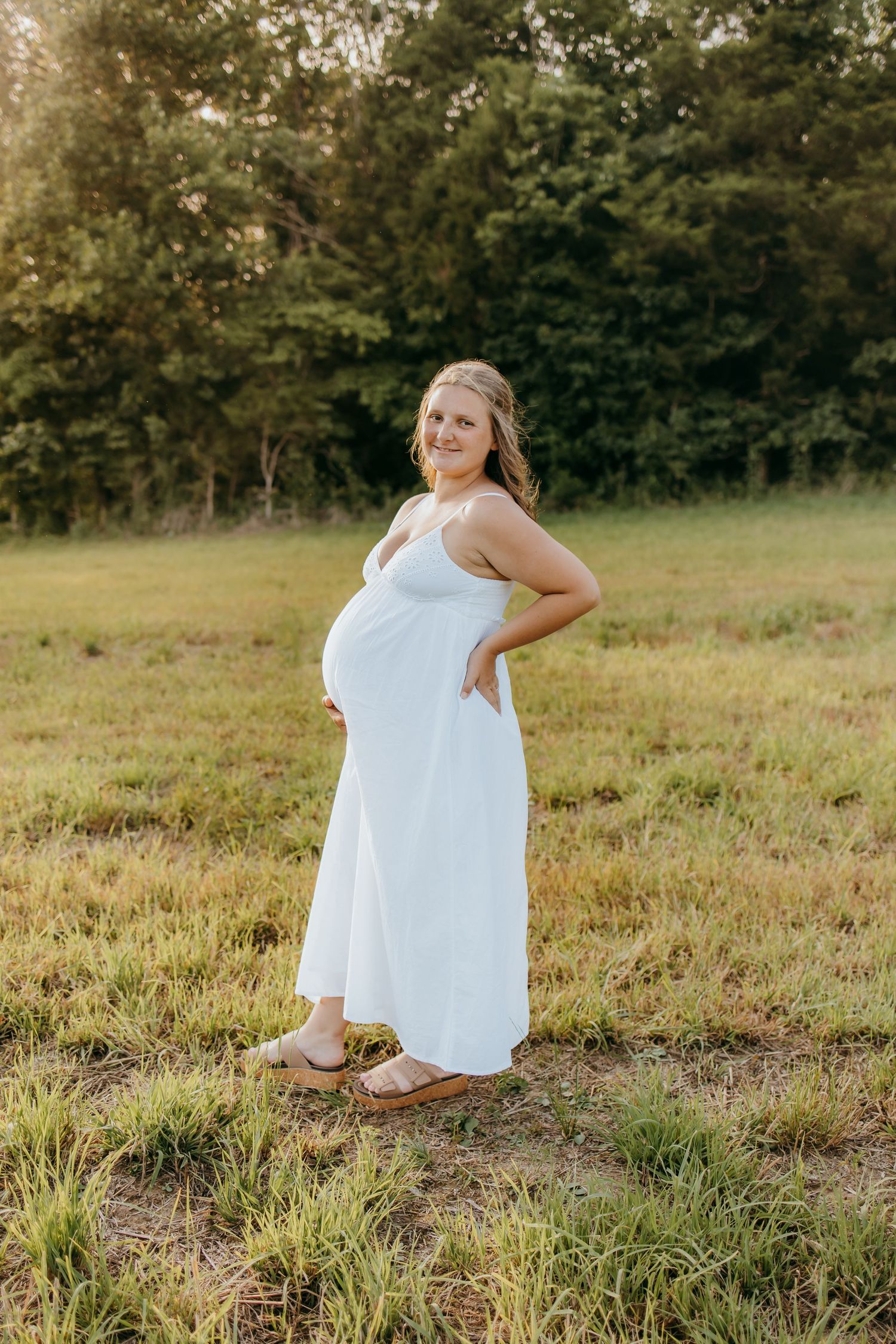 Maternity session in white maxi dress posing in a sunny green field during a maternity photo session.