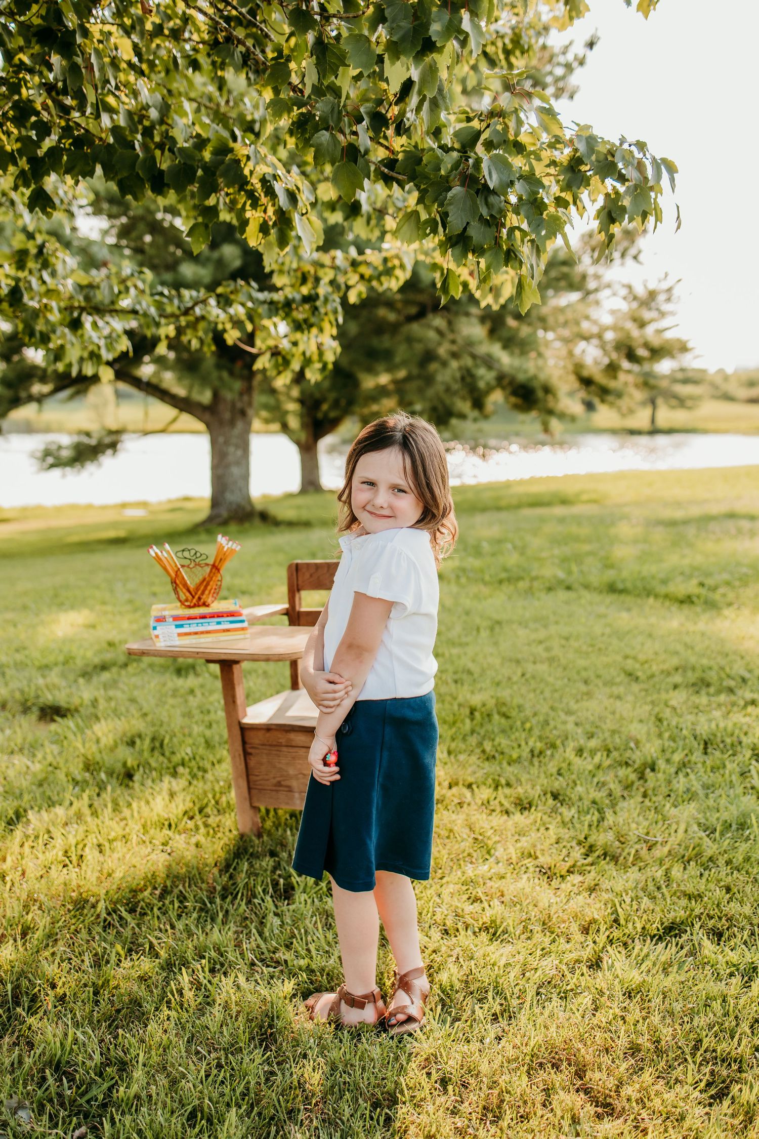 Young girl in white top and blue skirt stands on green grass near a wooden desk with school supplies outdoors.