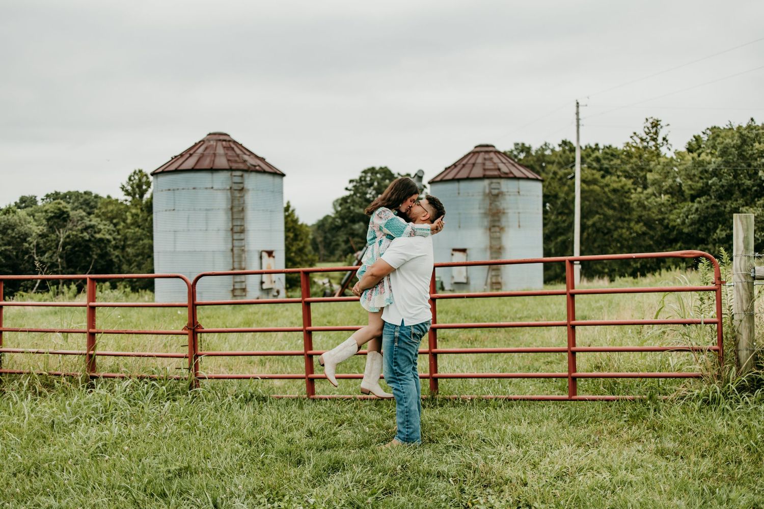 Couple embracing by red farm gate with grain silos in background on a cloudy day.