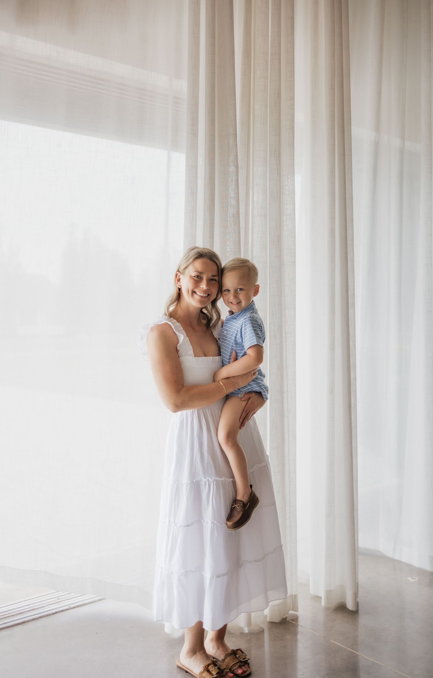 Woman in white maxi dress holding a young toddler boy, standing in front of sheer white curtains.