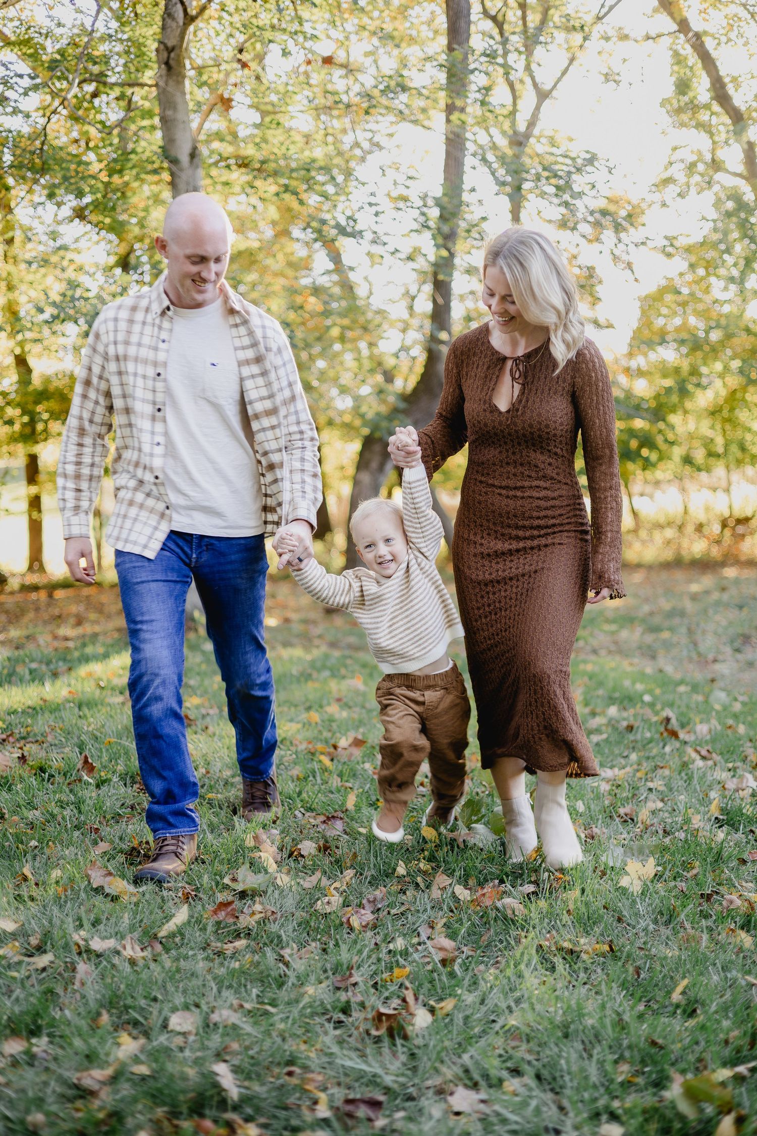A family of three walks through a sunlit autumn park, parents holding their toddler's hands between them.