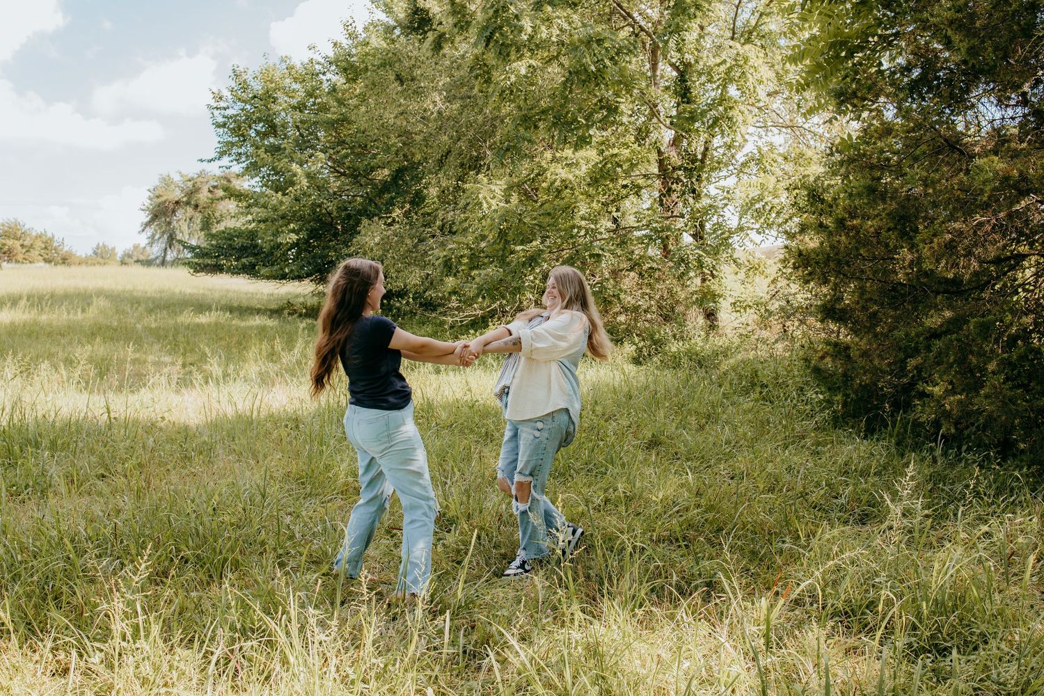 Two young women holding hands and dancing in a lush green meadow on a sunny day.