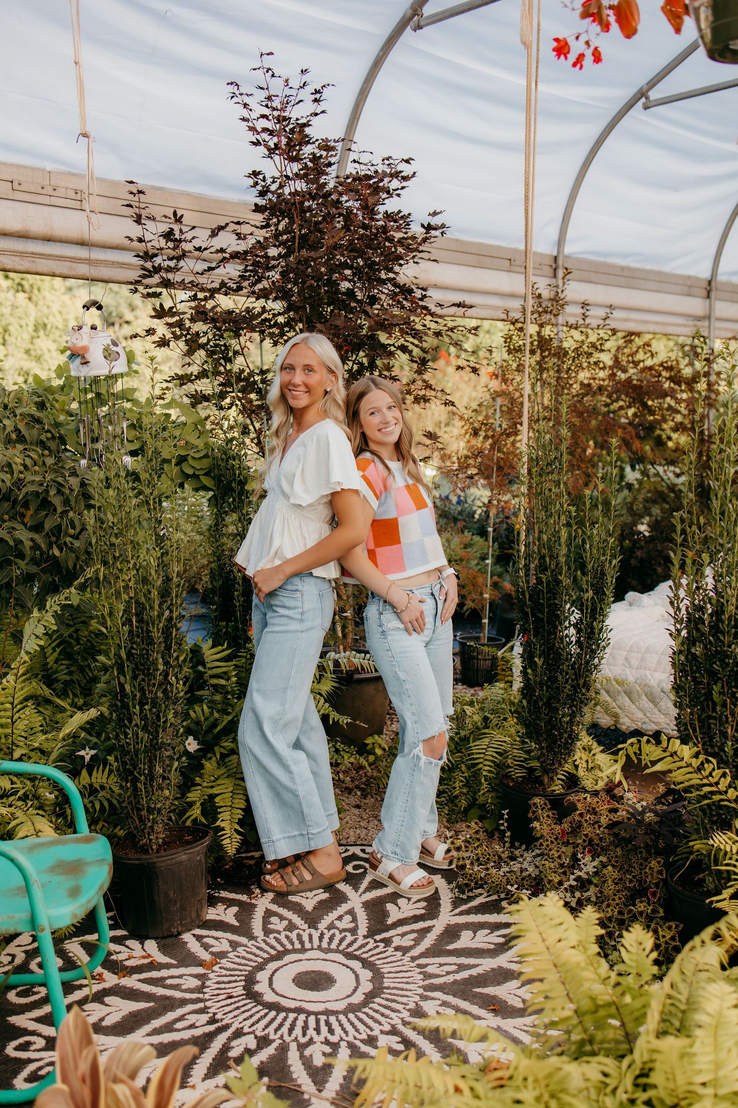 Two seniors in jeans pose back-to-back in a lush greenhouse surrounded by ferns and potted plants on a patterned rug.