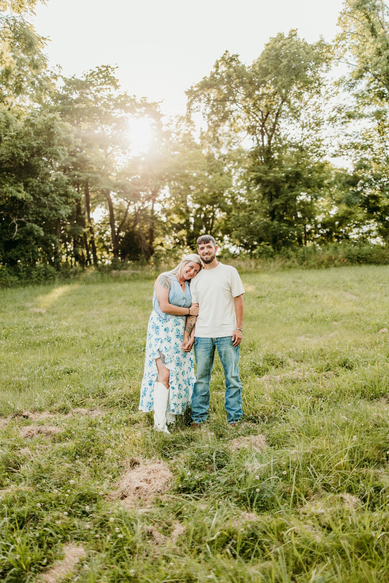Couple embracing in a sunlit green meadow, woman in floral dress leaning on man in jeans and white tee.