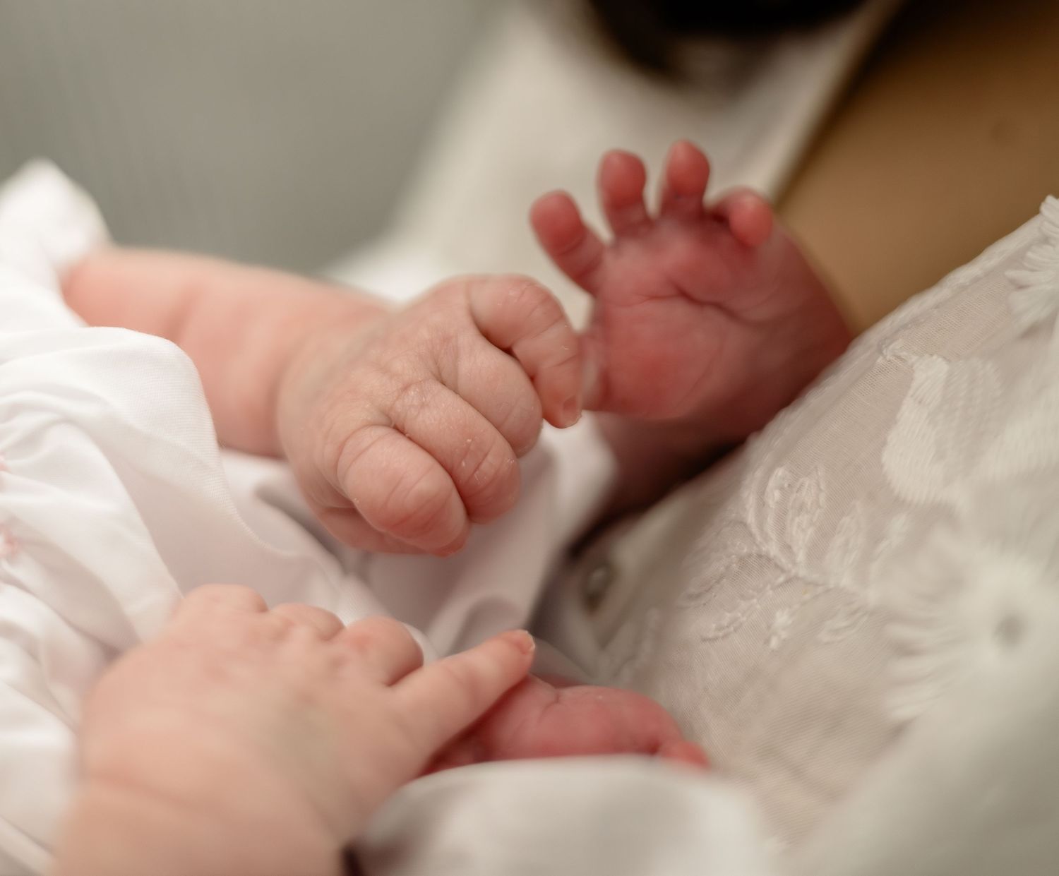Close-up of newborn baby hands grasping gently while dressed in a white christening gown.