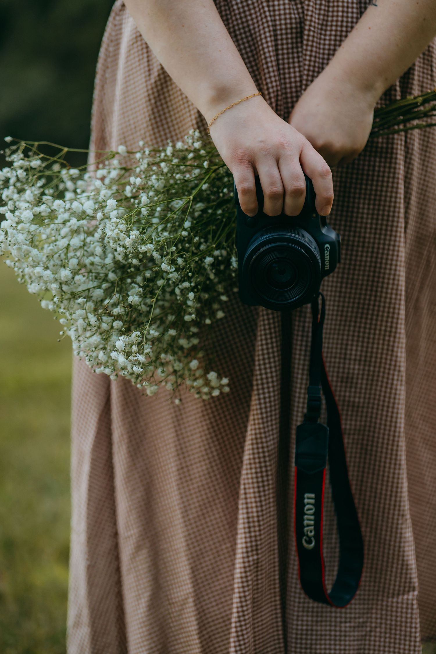 Woman in plaid dress holding Canon DSLR camera and white baby's breath flower bouquet outdoors.